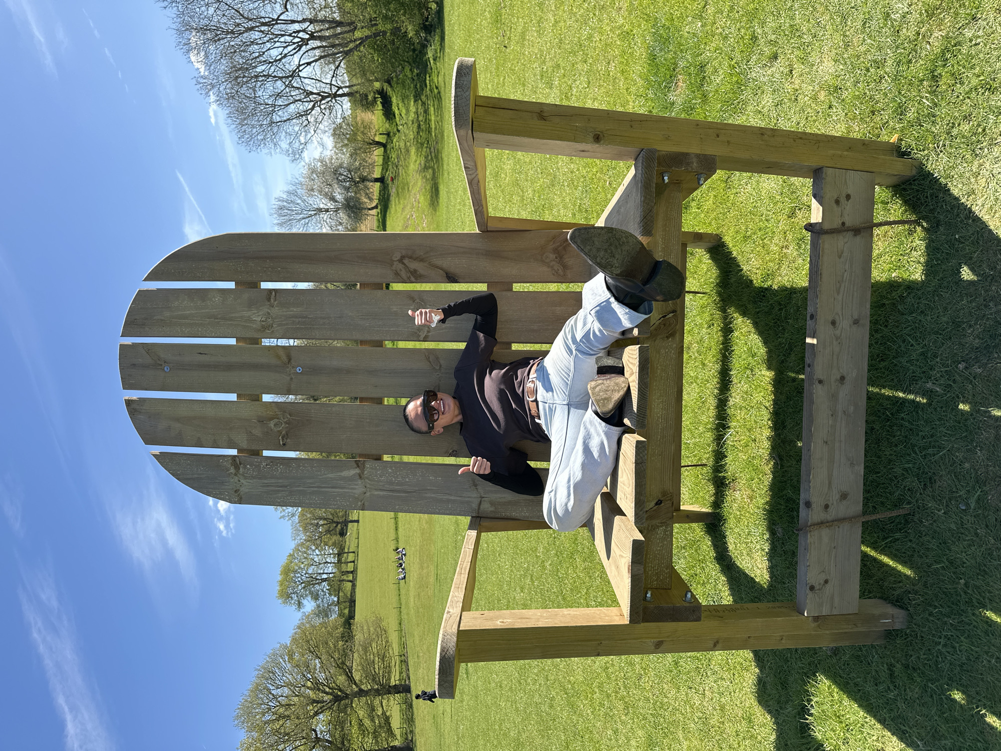 A medium shot, taken outdoors, features a woman lounging on an oversized wooden Adirondack chair set amidst a lush green lawn, against a backdrop of trees and a clear blue sky with wispy clouds, suggesting a rural setting.
The woman, clad in a dark long-sleeved shirt, light-colored jeans, and black boots, wears sunglasses and reclines casually in the chair, giving a thumbs-up gesture. Her positioning and expression suggest a relaxed and playful mood.
The Adirondack chair itself is constructed from unfinished wood. The expansive lawn, typical of a park or large garden, is well-maintained, and distant trees add depth to the scene.
While the exact location within the city of Sevenoaks, United Kingdom, is impossible to pinpoint, the presence of a large, grassy area suggests a park or expansive garden.