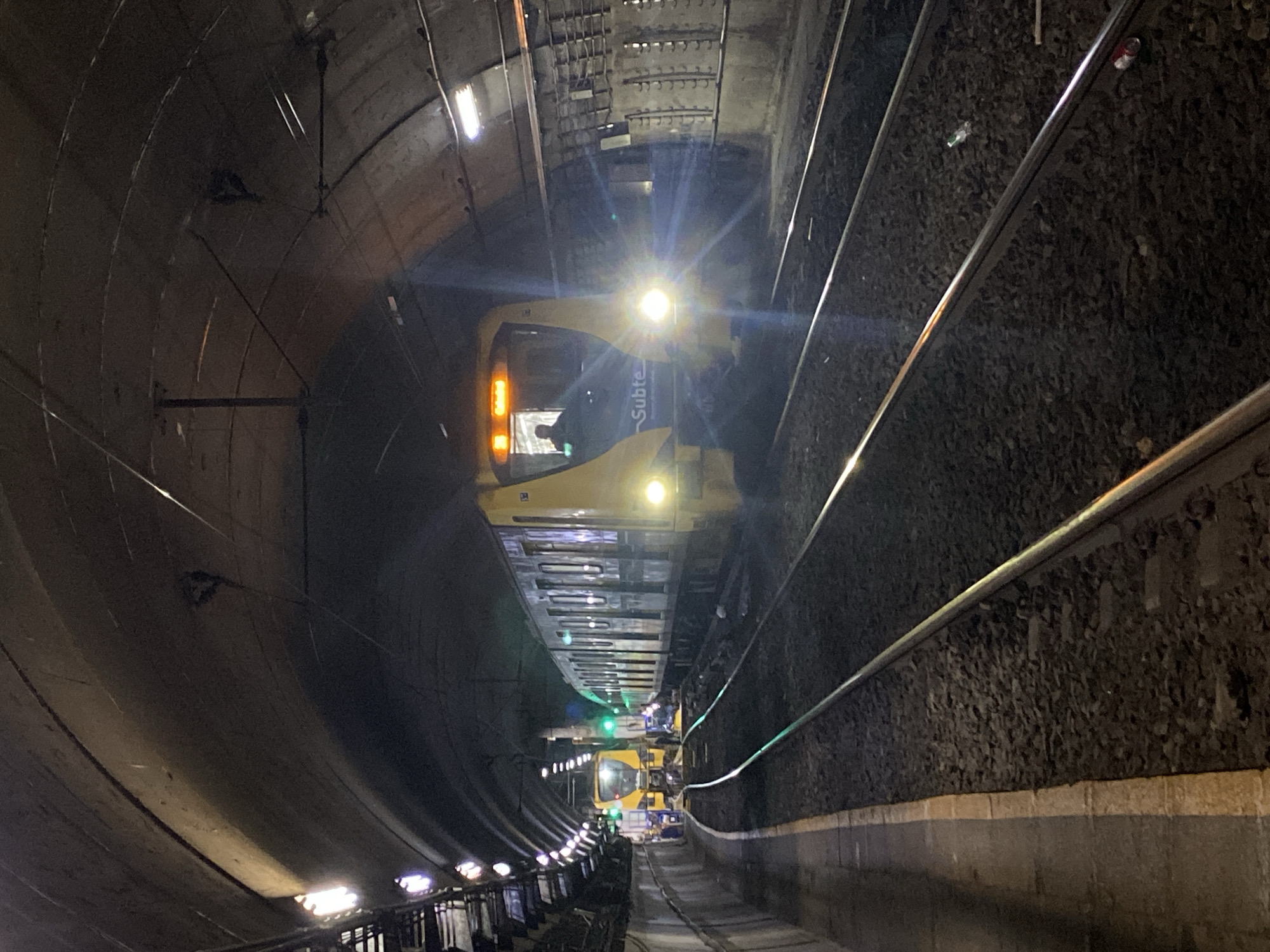 A subway train labeled "Subte" in yellow and white is traveling through a tunnel in Buenos Aires, Argentina. The tunnel is lined with concrete and features overhead lighting fixtures. The train's headlights are illuminated, and a person is visible in the operator's compartment. The subway tracks are visible on either side of the train, with ballast along the sides. In the distance, another train or maintenance vehicle is present.