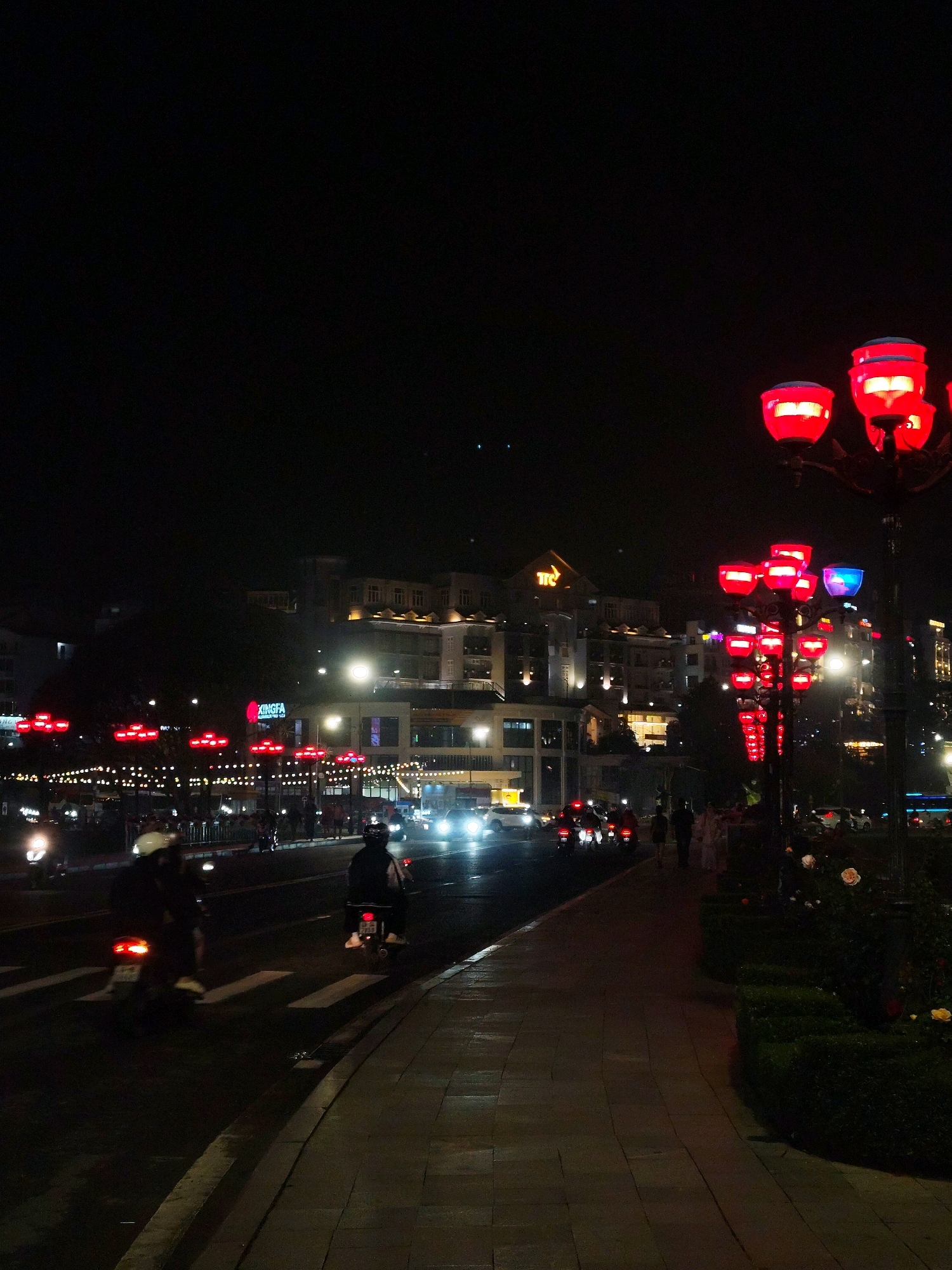 The image captures a nighttime scene on a street in Da Lat, Vietnam, characterized by vibrant red lanterns illuminating the surroundings. On the right side of the frame, a lamppost adorned with multiple red lanterns stands tall, casting a warm glow. Adjacent to it, a manicured green hedge with scattered roses adds a touch of nature to the urban landscape. The street itself is wet, suggesting recent rain, and lined with pedestrian walkways paved with stone tiles.
Several individuals are present, most notably riding motorcycles. Two motorcycles are prominent in the foreground, with riders wearing helmets and moving across a pedestrian crossing. Further down the street, more motorcycles and a few pedestrians can be observed.
The background reveals a cityscape with buildings of varying heights, many of which are lit up, creating a lively atmosphere. A prominent sign for "KINGFA" is visible on one of the buildings, indicating commercial activity. The overall impression is of a bustling city at night, enhanced by the decorative lighting.
