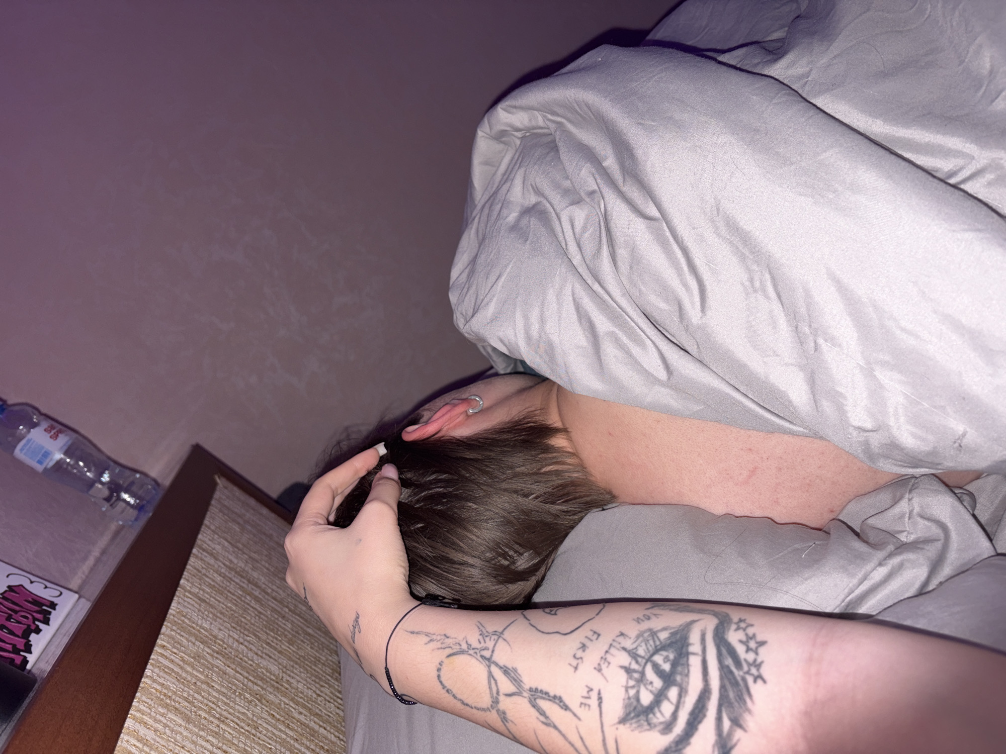 The image depicts an intimate bedroom scene in the city of Chaikovsky, Russia. Two individuals are partially visible in a bed under dim, warm lighting.
In the foreground, the left arm and hand of one individual are prominent. The arm is extensively tattooed, featuring a large eye design with radiating lines, three stars beneath it, and text that partially reads "YOU KILLED ME FIRST". A small script tattoo, possibly "always", is visible on the wrist, along with a bracelet made of dark beads. The hand, with white-tipped fingernails, is resting on the head of a second individual.
The second individual is seen from the back, displaying brown, short-to-medium length, disheveled hair. Their left ear is visible, adorned with two silver hoop earrings. The exposed skin of the neck and upper back shows a fair complexion with some minor redness. This individual is lying on their side, mostly covered by a light grey duvet or blanket.
In the upper left portion of the image, a wooden bedside table with a textured light-colored panel is visible. On the table, a clear plastic water bottle, labeled "SPRING", and a white object with pink lettering are present. The background wall is a plain light purple color.