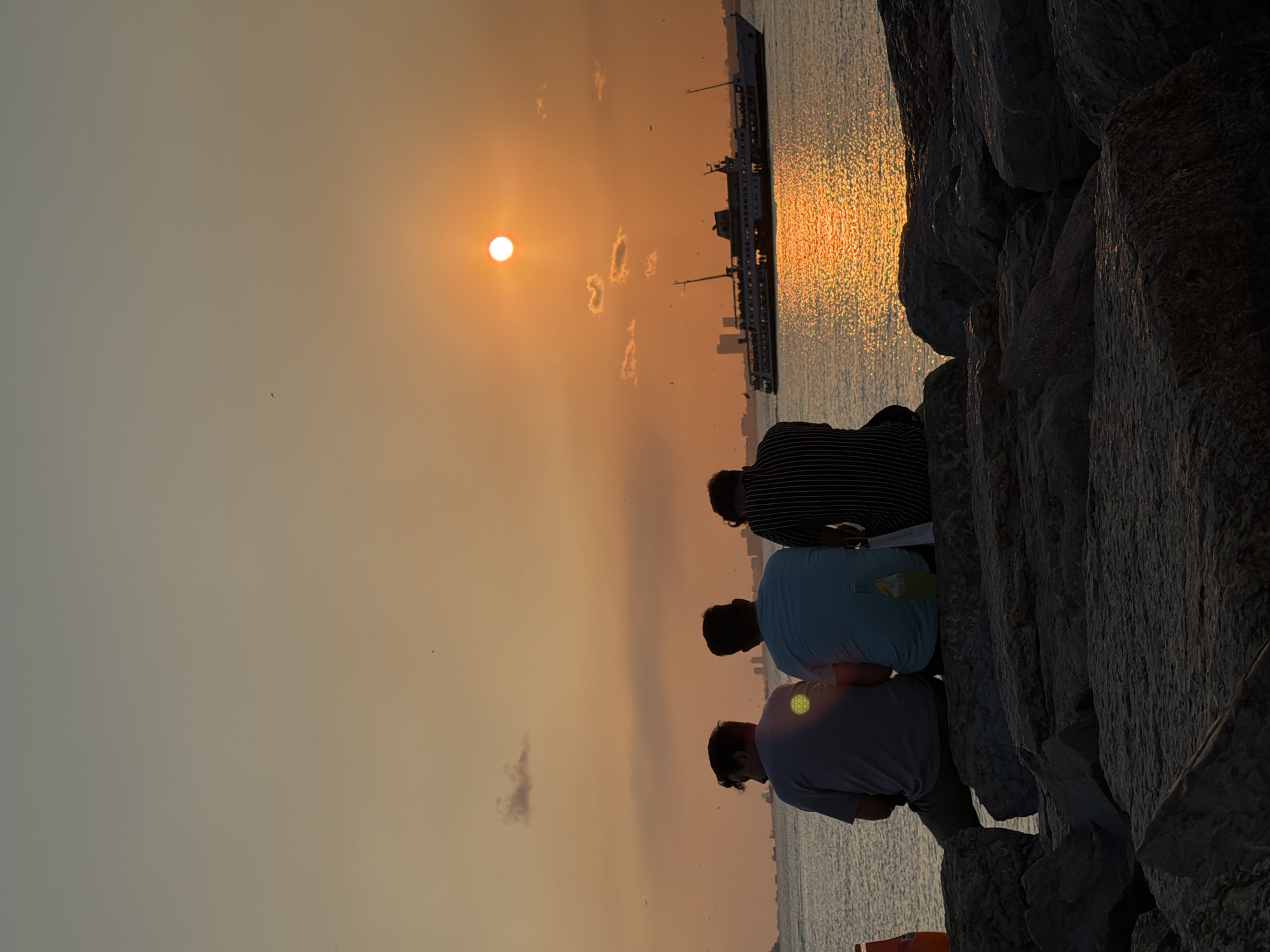 Three individuals are seated on large rocks facing a body of water during sunset. The sky is a gradient of orange and yellow, with the sun positioned high and to the right, casting a reflection on the water. A large ferry is visible on the water, moving from right to left, with a city skyline in the distance. One person is wearing a grey t-shirt and olive-colored pants, another a light blue t-shirt, and the third a black and white striped shirt. A yellow-green bottle is placed next to the person in the light blue t-shirt, and an orange cone is partially visible on the left. The rocks appear to be a breakwater or pier, typical of the coastline in Kadıköy, Turkey, where such waterfront views and ferry services are common.