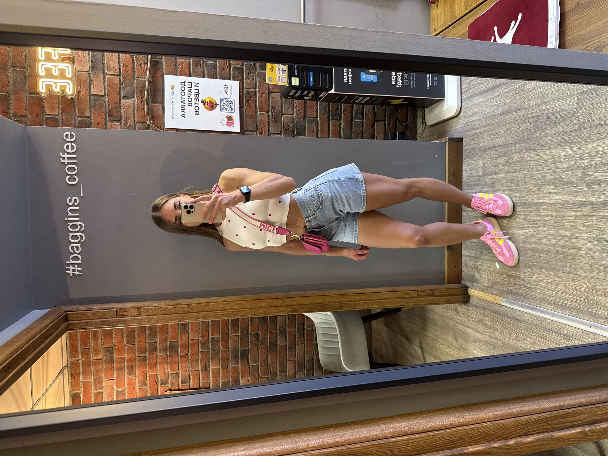A young woman is taking a mirror selfie in what appears to be a coffee shop in Saint Petersburg, Russia. She is wearing a white crop top with red hearts, light-wash denim shorts, and pink sneakers. A small pink crossbody bag is slung over her shoulder. The background shows a brick wall with a neon sign that reads "333" and the hashtag "#baggins_coffee." A large mirror reflects the woman and the interior of the establishment, including a doorway, a chair, and a charging station. The floor is made of light-colored wood planks.