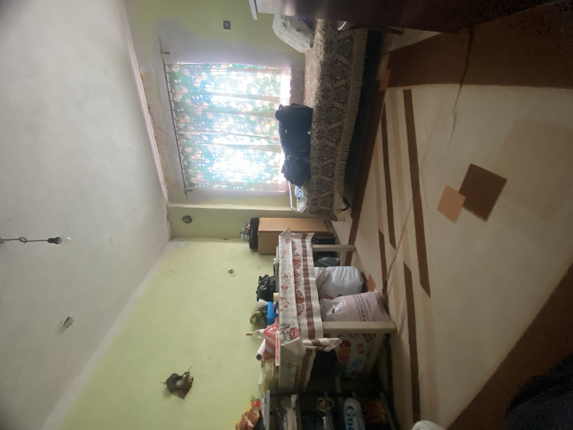 The image shows an interior scene, possibly in Katuntsi, Bulgaria. The walls are painted in light green and white tones. A window is partially visible, covered with floral-patterned curtains. Adjacent to the window is a bed covered with a decorative bedspread. There are some bags placed by the window. To the right of the frame, there is a carpet with geometric patterns. In the foreground, there's a cluttered shelving unit stacked with bags, containers, and bottles. A small table or stand is draped with a cloth, also containing various objects. The lighting seems bright near the window, casting shadows in other parts of the room. The room appears cluttered and somewhat disorganized. There are no people visible.