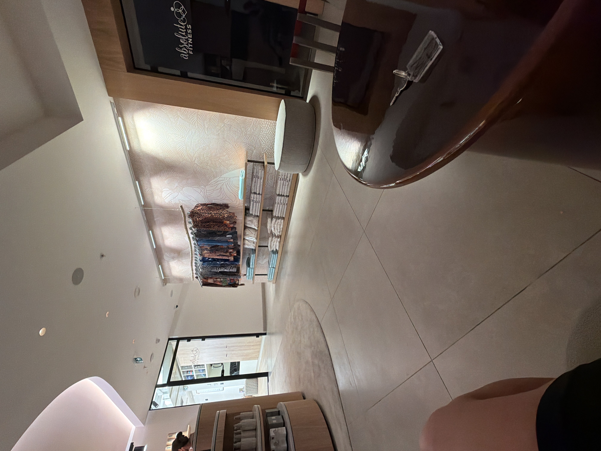 The image depicts the interior of a retail establishment, likely a boutique or studio, with a modern and clean aesthetic. The flooring is tiled in a light neutral tone. In the foreground, a dark, highly polished circular table is visible, with a set of keys resting on its surface. Further into the scene, to the left, is a curved, multi-tiered display unit made of light wood, featuring shelves stocked with bottles and other products. Adjacent to this, a person's leg is visible, suggesting they are seated.
The middle ground of the image showcases a display of clothing hanging on a rack, featuring various patterns and colors. Below the clothing, shelves are neatly stacked with folded items, possibly towels or apparel. A light grey, cylindrical ottoman or stool is positioned in front of a wall with a textured, patterned wallpaper, illuminated by track lighting.
To the right of the clothing display, a large glass-fronted door or window is present, revealing a dark interior space. A logo is visible on the glass, reading "absolute FITNESS." The wall next to this entrance is paneled with warm-toned wood.
The overall environment appears to be a commercial space designed for retail or services, characterized by its contemporary design, ample lighting, and organized displays. The presence of the "absolute FITNESS" logo suggests a connection to a fitness brand or facility. The location is not specified in the image, and the city of Boreti, Montenegro is not visible or identifiable within the visual information provided.
