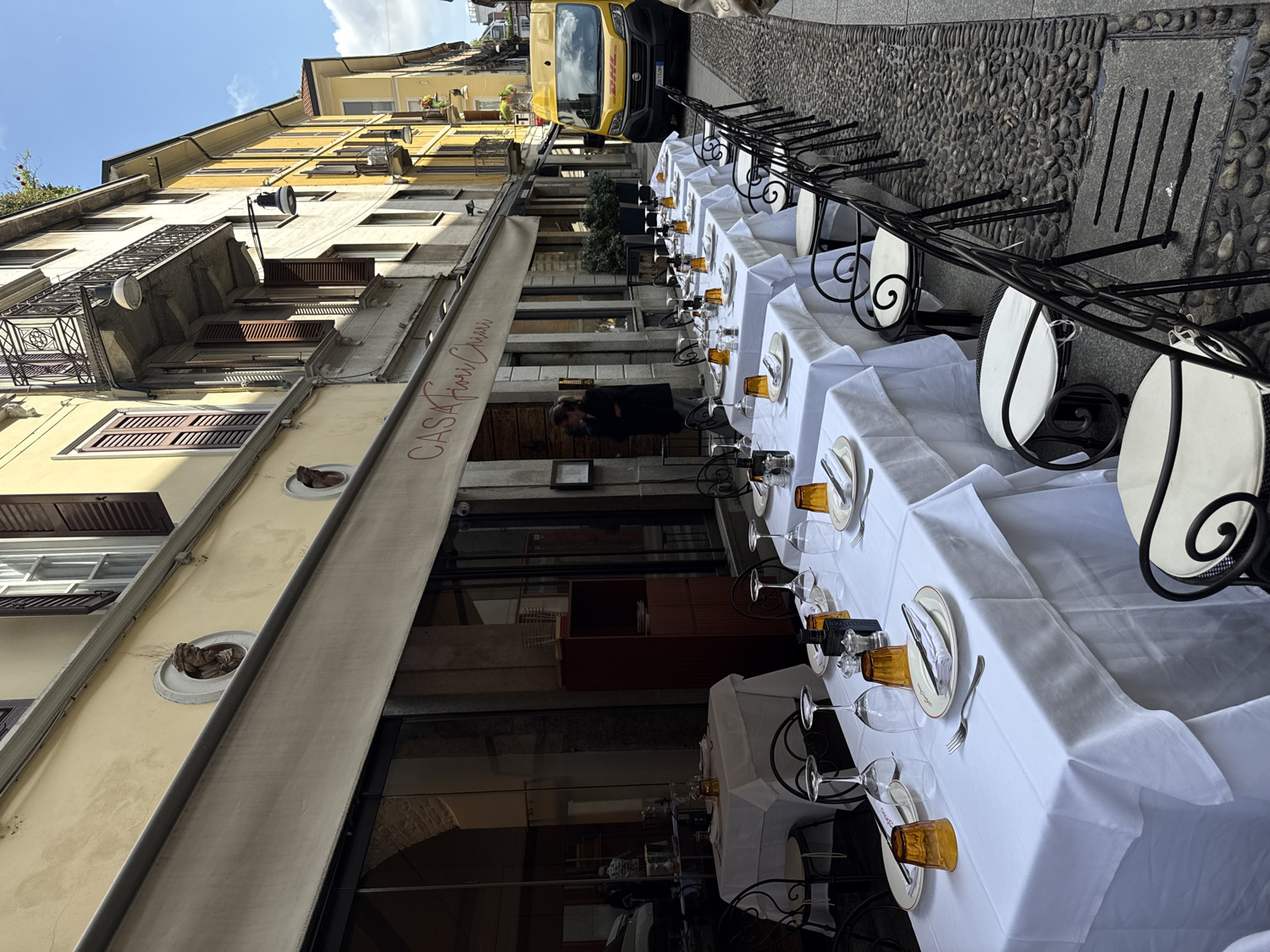 The image displays an inviting outdoor dining setup along a narrow, cobblestone street in Milan, Italy, captured from a slightly upward and tilted perspective.
Numerous tables are meticulously arranged along the right side of the street, each covered with a crisp white tablecloth. Every place setting includes a white plate, cutlery wrapped in a napkin, an amber-colored drinking glass, and a wine glass. Complementing the tables are elegant chairs with ornate black wrought iron frames and circular white or light-colored seats and backrests.
To the left, a historic multi-story building with a warm yellow facade rises. It features several windows fitted with brown wooden shutters, some open, some closed, and small balconies adorned with black iron railings. Wall-mounted lamps are visible on the building's exterior. A large, light-colored awning extends from the building over the dining area, providing shade. The name "CASA ANNA CHIESA" is clearly visible in red script on this awning.
The street itself is paved with rough cobblestones. Further down the street, a bright yellow DHL delivery van is parked, and its logo is clearly visible. A person wearing a dark top is partially visible on the right side, appearing to be a staff member engaged in setting up or organizing the outdoor dining area.
The scene is brightly lit by natural daylight under a clear sky with some clouds, suggesting it is mid-day or early afternoon. The overall ambiance is one of readiness and quiet anticipation before meal service.
