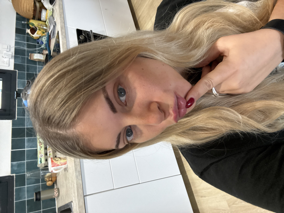A female subject with long, parted blonde hair, blue eyes, and visible freckles is captured in a head-and-shoulders frame. She is wearing a black top. Her lips are pursed forward, and her right index finger, adorned with a silver ring and dark red nail polish, is positioned against her lower lip. She is looking directly into the camera.
The scene is a modern kitchen located in the city of Rousset, France. The background features a backsplash of dark teal square tiles with white grout. Below this are light-colored countertops and white, handleless lower cabinets. On the left side of the counter, a stainless steel sink is partially visible, alongside a dark insulated bottle, a clear glass bottle, a white mug with a decorative image, and a carton of eggs. A dark-framed window is present above this section. On the right side of the counter, a black induction cooktop is visible, with a dark oven or microwave integrated into the white cabinetry below. Additional objects, including a woven basket and various jars, are arranged on the counter. A white electrical outlet with cords is mounted on the tiled wall. The floor, visible at the bottom of the frame, is a light wood-look material.