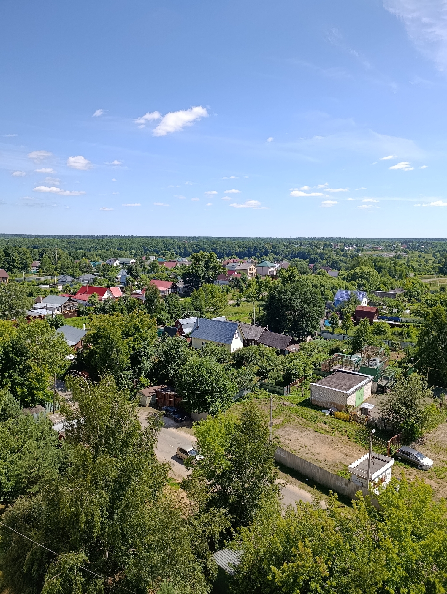 This is a high-angle, daylight, full shot of a semi-rural residential area in Alexandrov, Russia. The scene is dominated by lush green trees and vegetation interspersed with numerous houses with various roof colors, including red, gray, and blue. A few roads are visible, one of which has a silver car parked on it and another with a light-colored car driving away. Several small outbuildings and fences are also present. The sky is a clear blue with scattered white clouds. The overall impression is of a tranquil, well-treed community under a bright sky.