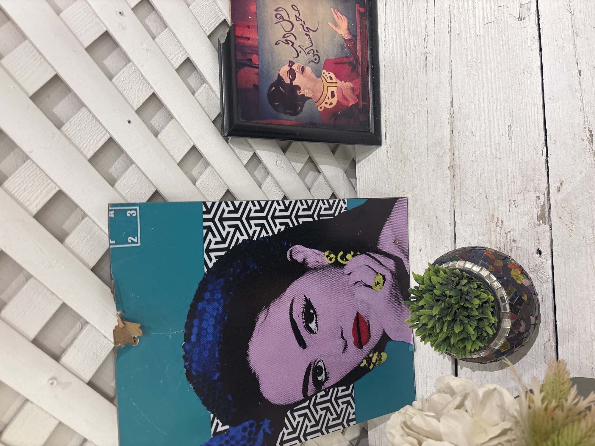 The image depicts a close-up of a wall adorned with two pieces of art and a small potted plant. The wall itself is constructed of white wooden lattice work.
To the left, a large, rectangular artwork features a pop-art style portrait of a woman with purple skin, dark hair, and bold red lips. She is wearing large, yellow hoop earrings and a ring. Behind her head, a blue element with a sequined texture is visible, and to her right, a geometric black and white pattern. A small, torn section of what appears to be cardboard is visible at the top edge of this artwork, suggesting it might be a record sleeve. Numbers "2" and "3" are faintly visible on the upper right corner of this artwork.
To the right of the large artwork is a smaller, framed picture. This frame is black and contains an image of a woman in a red dress with elaborate gold trim around the neckline. She has her hair styled up, is wearing sunglasses, and is speaking with her hands gesturing. Arabic script is present on the right side of this artwork.
In the foreground, resting on a white wooden surface, is a small, round planter made of mosaic tiles in various colors. It contains a vibrant green, artificial plant. To the far left, out of focus, is a large, white floral arrangement.
The overall scene suggests a decorative display, possibly within a home or a shop. There is no information in the image to confirm or deny a location in the city of Amman, Jordan.