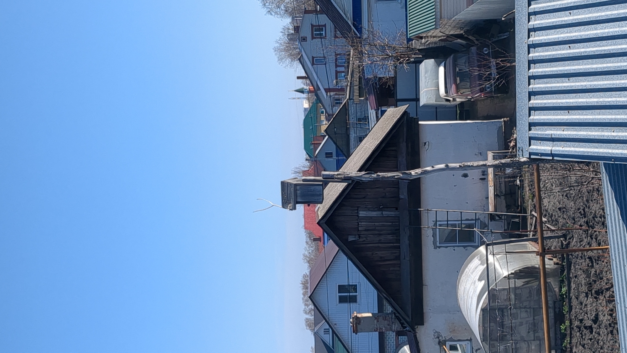 The image presents an outdoor scene featuring a residential area under a clear, bright blue sky. The perspective is slightly elevated, looking across an array of buildings.
In the foreground, on the right side, a corrugated metal fence or wall suggests an enclosure or part of an industrial-style structure. Adjacent to this, in the midground, is a rustic wooden building with a dark, steeply pitched roof. A small, dark, box-like structure, possibly a birdhouse or a ventilation cap, sits on its roof, from which a distinctive white, Y-shaped twig extends. To the right and slightly below this wooden building, a small greenhouse or cold frame with a translucent, arched roof is visible. Further right, a white building wall with a window and a large, light-colored cylindrical tank or container are present. A dark red car is partially seen behind some bare branches, nestled among the structures.
The background reveals a collection of houses with diverse architectural styles and roof colors, including red, green, and various shades of grey and blue. Some houses feature light-colored siding, while others are darker. Bare trees are interspersed throughout the landscape, indicating late winter or early spring. A slender, pointed spire or antenna is faintly visible in the distant horizon.
The sky is a uniform, vivid blue, suggesting a sunny day with no clouds. There are no people or discernible activities taking place. The scene projects a quiet, possibly suburban or semi-rural residential atmosphere. No visible text is present in the image. The environment is consistent with a residential setting in Ulyanovsk, Russia.