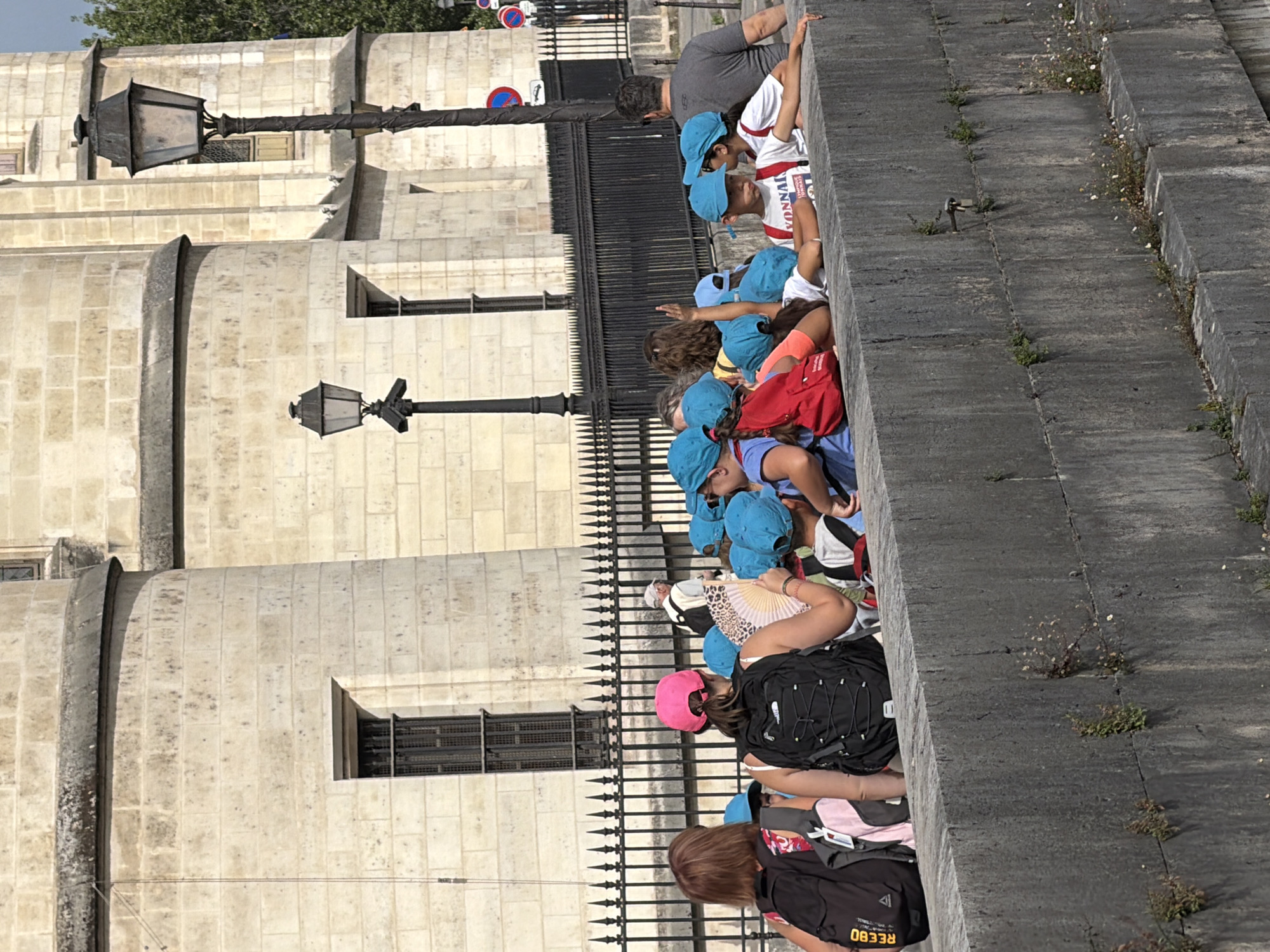 A group of children wearing blue hats and some adults are gathered along a stone wall in Paris, France, likely near a historic building. The children appear to be on a field trip, as many are wearing backpacks and some hold a fan. In the background, a large stone building with windows and a lamppost are visible. The scene suggests a sunny day, with shadows cast by the buildings and people.