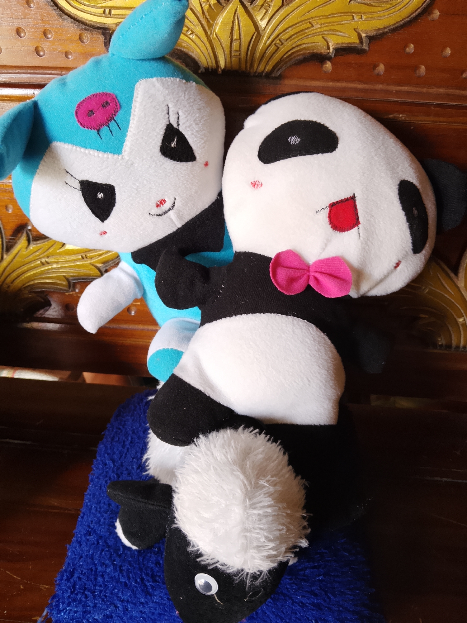 Two stuffed toys, a blue and white creature and a panda, are positioned on a wooden surface. The blue and white toy has a pink stitched design on its head and black eyes with red stitching accents. The panda toy is wearing a pink bow tie and has black eye patches, red stitched accents on its face, and is resting on a blue fuzzy material. The background features ornate, carved wooden furniture. This image was taken in Kasiman, Indonesia.