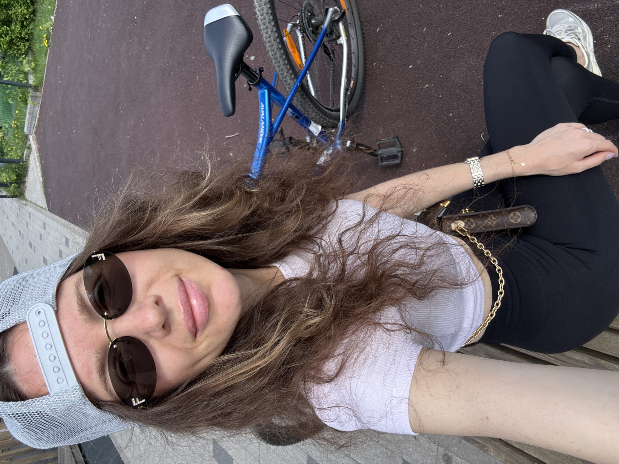 A selfie shows a woman sitting on a park bench, wearing sunglasses and a baseball cap. She is in black leggings and a white t-shirt, with a purse with a gold chain. A blue bicycle lies on the ground next to her. The location is a park in Moscow, Russia, based on visual cues in the image.