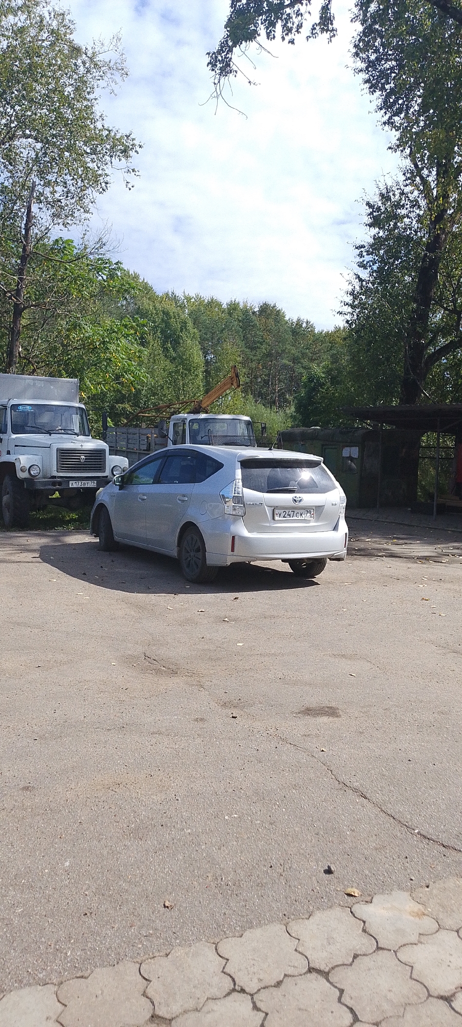 The image depicts an outdoor scene in Bira, Russia, focusing on parked vehicles and surrounding vegetation. In the foreground, a silver Toyota Prius is parked on an asphalt surface. The car's rear is visible, showing its license plate which reads "Y247CK 79". To the left of the Prius, a white truck with a large cargo box is parked, displaying a license plate that appears to say "A173BT 79". Behind these vehicles, a wooded area with dense green trees forms the background. A yellow crane is visible between the two trucks, suggesting a possible construction or industrial area. The sky above is partly cloudy with a bright, daytime light. The ground is a mix of cracked asphalt and decorative interlocking pavers in the lower right corner.
