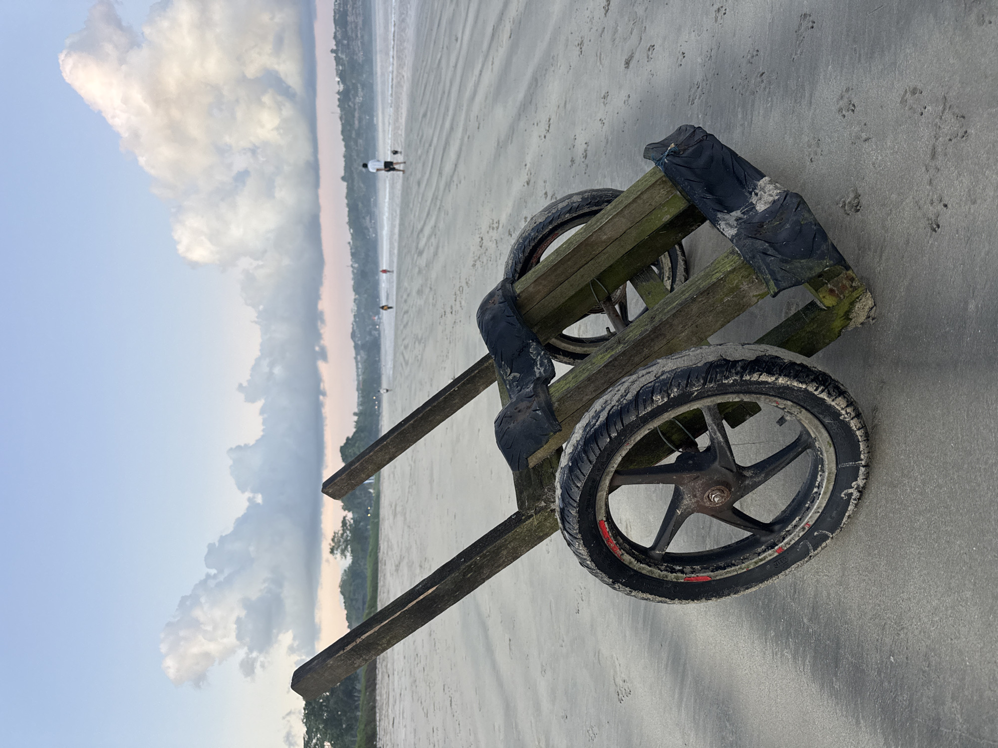 The image captures a wooden cart with two black wheels resting on a sandy beach. The cart has a rudimentary design, with visible wear and tear, including some black material wrapped around the upper part. The tires show evidence of sand and use. In the background, a beach scene unfolds with a few people visible, suggesting an interaction with the environment. The sky is partially cloudy. The location is the beach of Kuta Selatan, Indonesia.