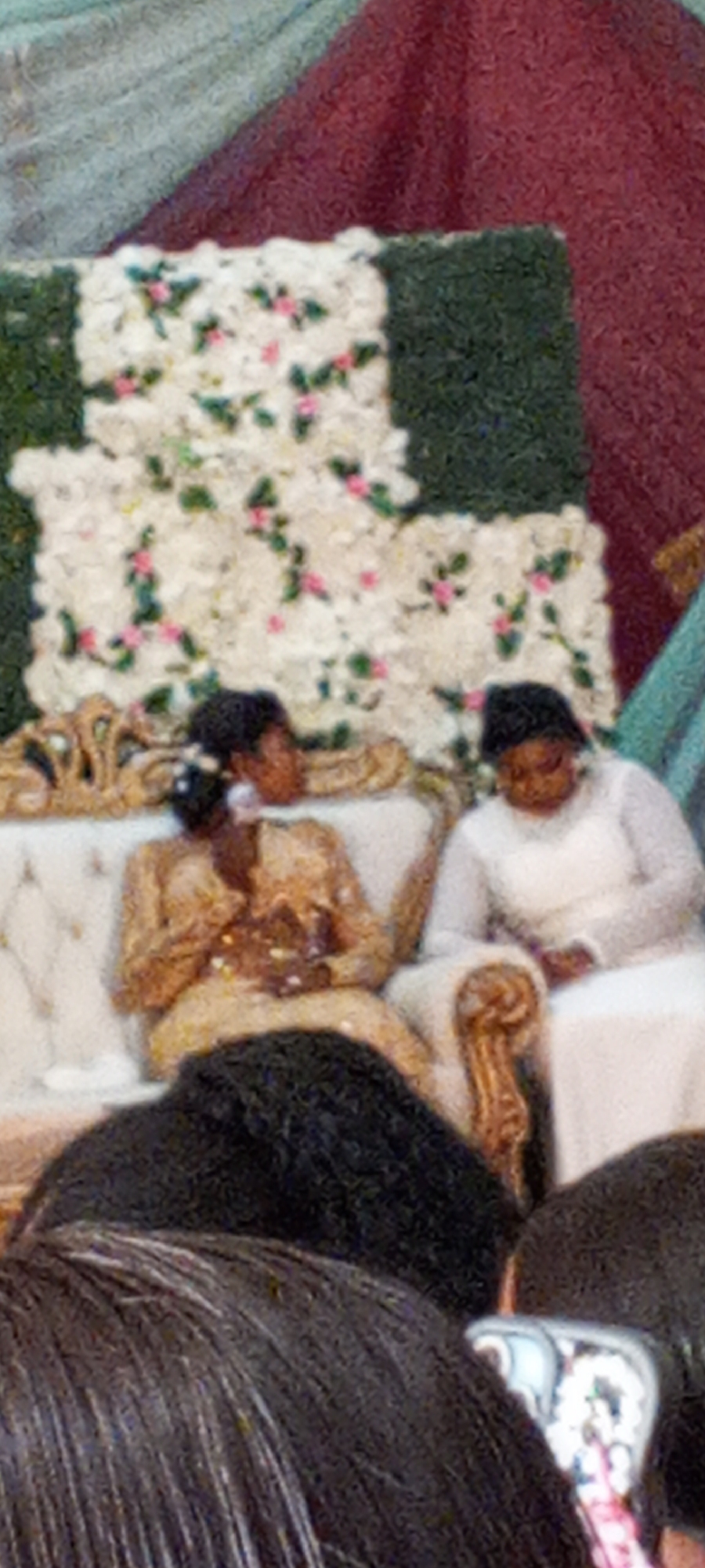 Two individuals are seated on a decorative couch in front of a floral backdrop in an indoor setting in Unknown, Unknown. The person on the left is wearing a gold dress and the person on the right is wearing a white dress. The floral backdrop is comprised of white flowers with green foliage and pink accents. The image is partially obscured by the heads of individuals in the foreground.
