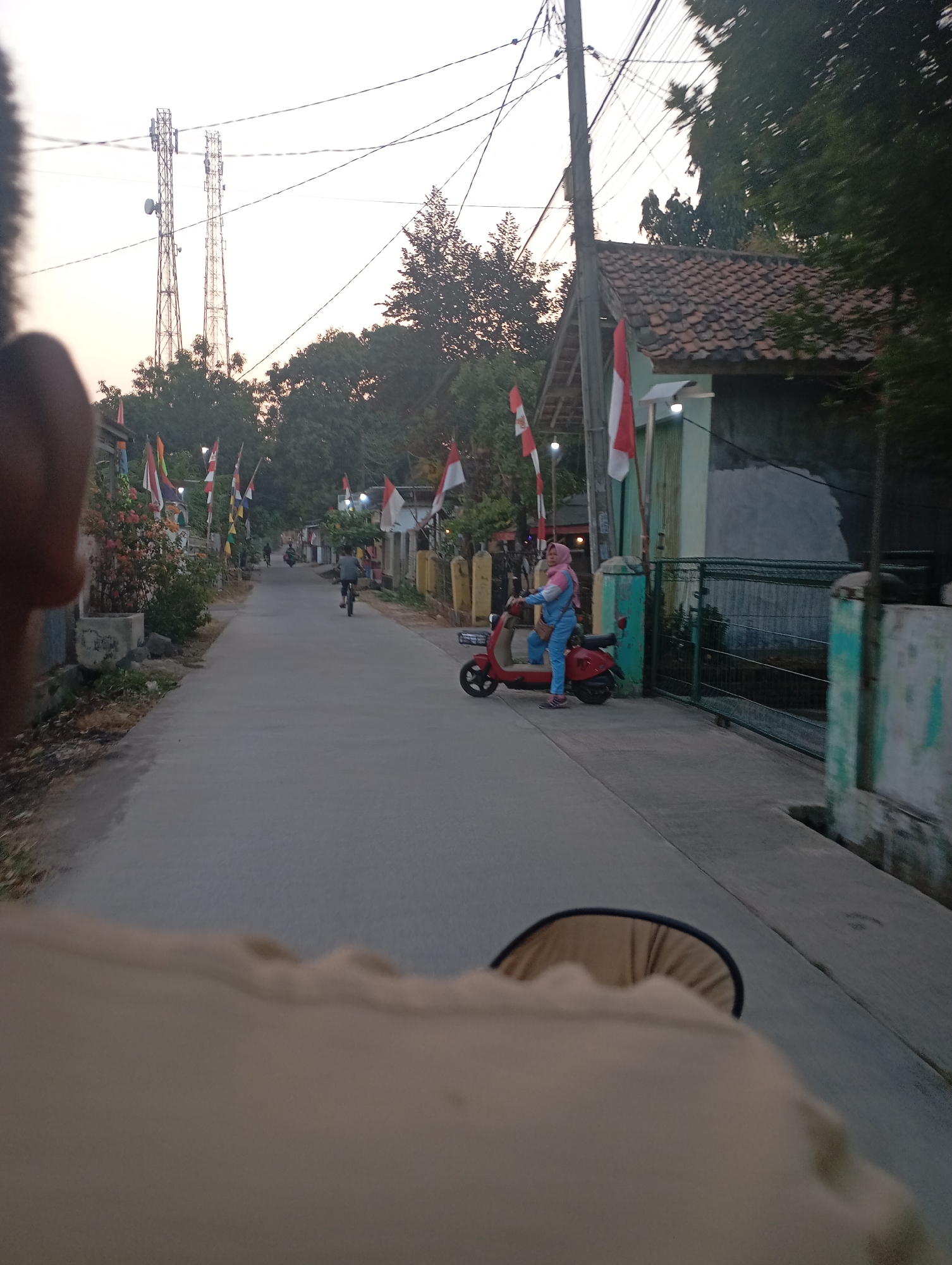 The image shows a street scene in Kecamatan Haurgeulis, Indonesia, during what appears to be late afternoon or early evening. In the foreground, the shoulder and head of the person taking the photo are visible, obscuring part of the scene. The street is paved and slopes slightly downwards. On the right side of the street, a red scooter is parked, with a woman wearing a pink hijab and blue and pink clothing sitting on it. She is looking towards the left. Adjacent to the scooter is a green metal fence. Further down the street, there are houses with tiled roofs, some adorned with Indonesian flags. On the left side of the street, there are more houses, plants, and a concrete planter. In the distance, two tall communication towers are visible against a pale sky, and several smaller flags are mounted on poles along the roadside. A person is riding a bicycle further up the street, and another motorbike is also visible. The overall impression is of a quiet residential street, possibly decorated for a celebration.