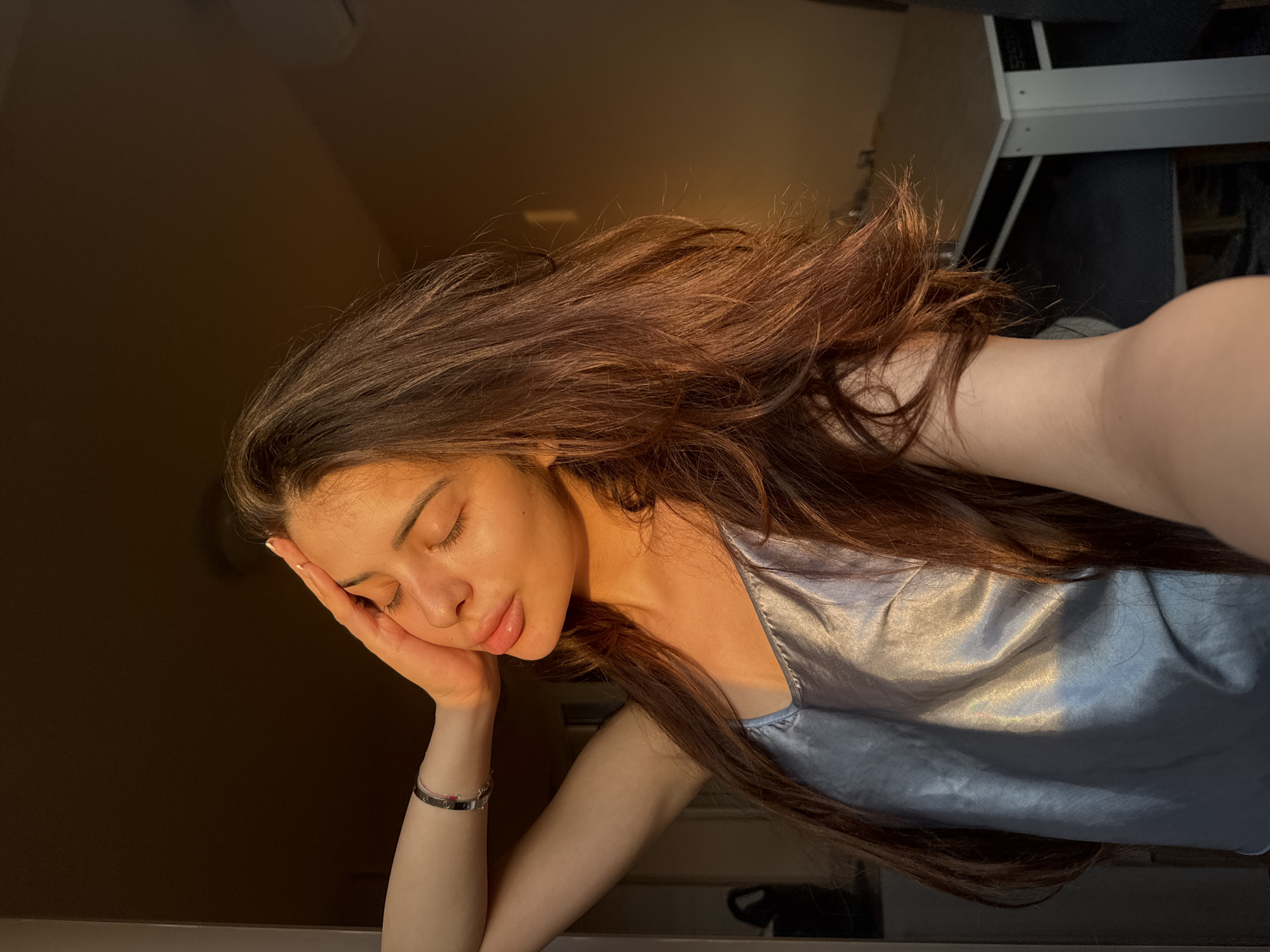 The image captures a young adult female indoors, located in the city of Plovdiv, Bulgaria, illuminated by a warm, golden light consistent with direct sunlight from a low angle, such as sunrise or sunset. The subject is fair-skinned, with long, wavy, dark brown hair cascading over her shoulders and chest. Her eyes are closed, and her head is tilted slightly upward and to the left. Her right hand is raised, with fingers resting gently on her forehead near her right temple. Her lips appear full. She is wearing a light-colored, possibly silver or light blue, satin or silk camisole top which exhibits strong highlights from the direct illumination. On her left wrist, a metallic, silver-toned bracelet with dark embellishments (possibly screws or engravings) is visible, layered above a thinner, light-colored bracelet. The background features a dimly lit interior. To the far left, a vertical light-colored architectural element is present. In the mid-ground right, a segment of a white table with white legs and dark chairs is discernible. An obscured light-colored object, possibly a wall-mounted unit, is visible in the upper right quadrant. The scene is characterized by strong contrasts between the brightly lit areas and the muted, shadowed portions of the room.