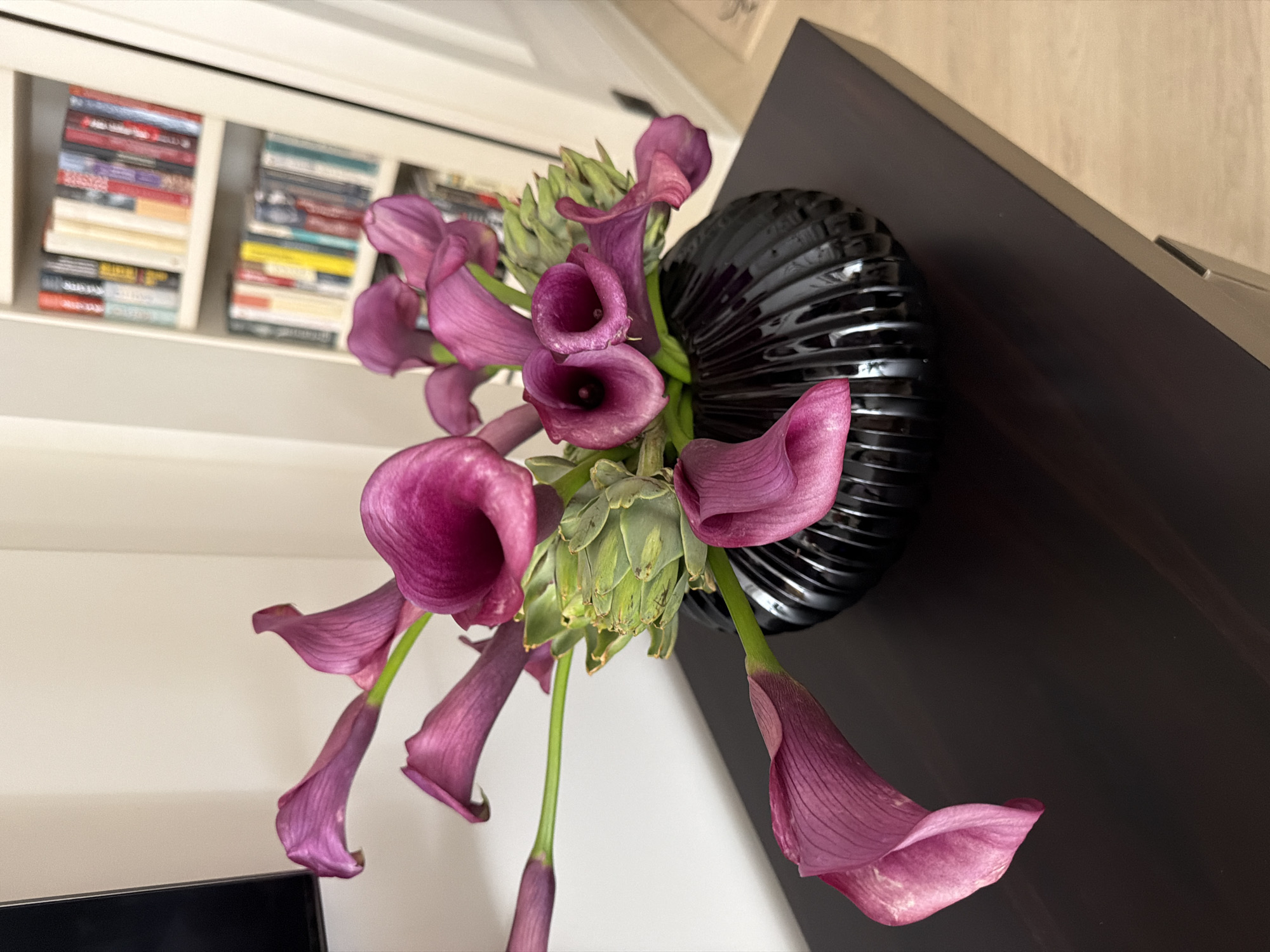 A floral arrangement of dark pink calla lilies and green artichokes sits in a ribbed black vase on a dark brown wooden table. Behind the arrangement, a white bookshelf is filled with colorful books. The background is a plain white wall. The scene is indoors.