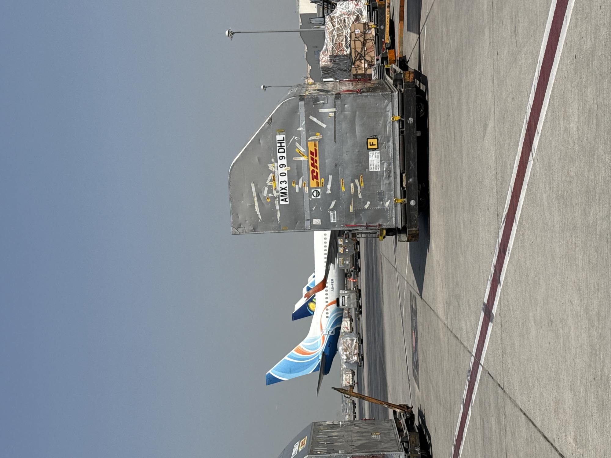 The image depicts an airport tarmac scene on a clear day, likely in Dubai, United Arab Emirates, given the presence of DHL cargo containers and an aircraft with an livery consistent with airlines operating in the region. In the foreground, a large, silver cargo container is prominently displayed, bearing the DHL logo and the identification code "AMX 3 0 9 9 DHL". The container is loaded onto a wheeled cart. Behind it, another similar cargo container is partially visible on the left. In the mid-ground, an airplane with a distinctive blue and orange fin with a swooping design is parked, its tail visible. Another airplane with a similar livery is positioned behind it. Several other cargo containers and baggage carts are scattered across the tarmac. The ground is concrete, marked with a red and white striped line. In the background, airport infrastructure, including light poles and buildings, can be seen under a hazy sky. There are no people visible in the image. The scene is one of typical airport operations, focusing on cargo handling and aircraft readiness.