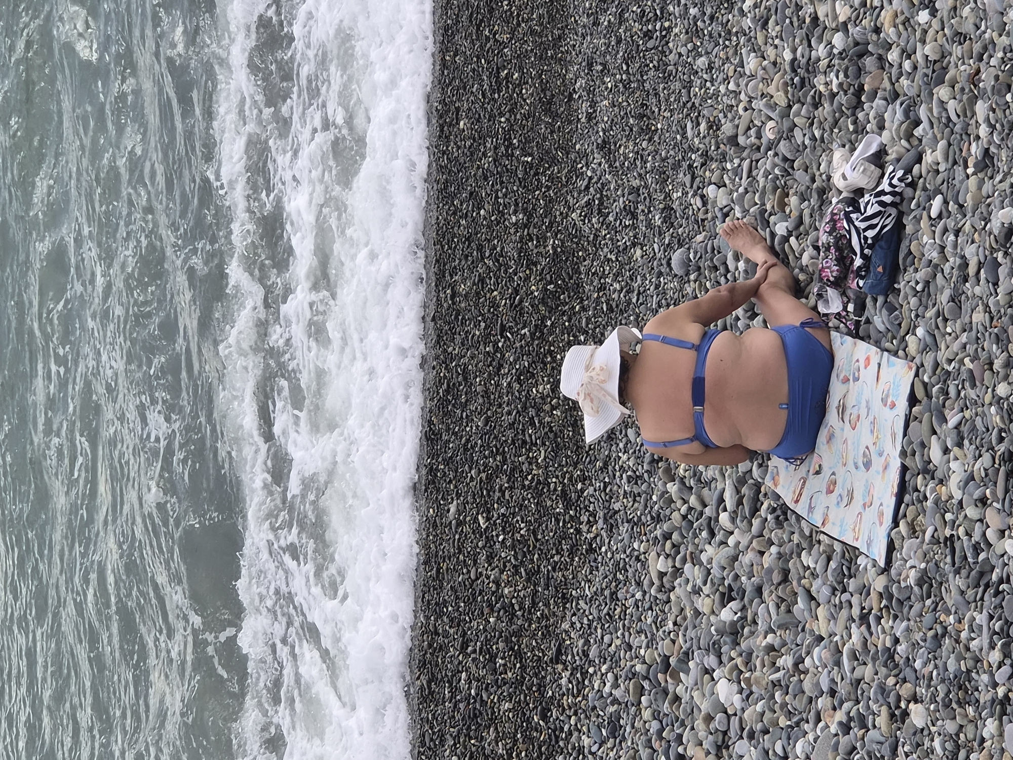 A woman wearing a blue bikini and a white hat lies on a colorful towel on a pebble beach in Sochi, Russia. The waves are crashing on the shore to her left. A pile of clothing and shoes is near her feet. The pebbles on the beach are mostly gray and black.