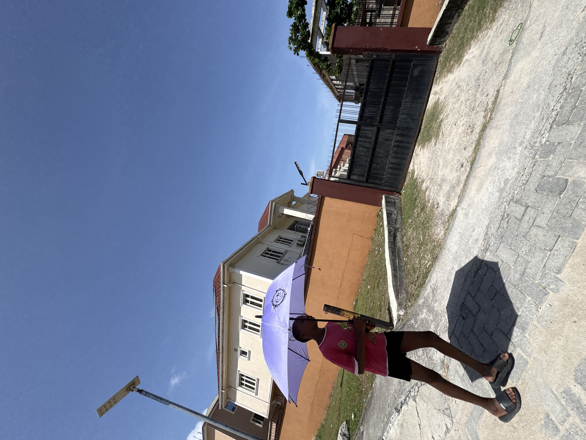 A young person walks on a paved area in Igboefon, Nigeria. They are holding a light purple umbrella. They are wearing a pink shirt, black shorts, and black sandals. Behind them is a building with beige walls and a red roof, a black metal gate, and a street light. The sky is blue and clear. The angle of the shot is slightly tilted.