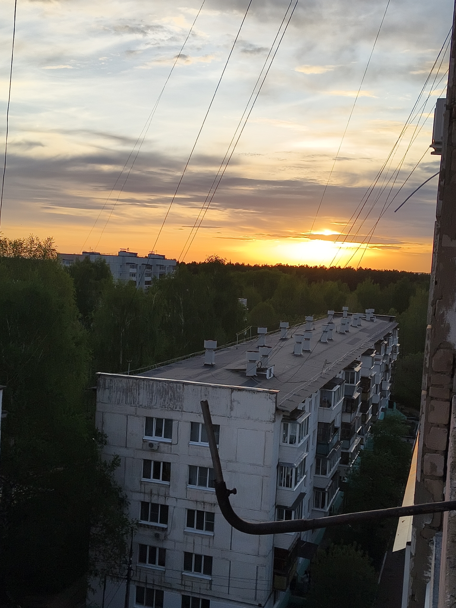 A high-angle view in an urban environment captures a sunset. Power lines stretch across the top of the frame against a sky with yellow-orange hues near the horizon and greyish clouds above. The middle ground features a dense collection of green trees, obscuring the ground. In the lower portion of the image, several buildings are visible. One building with a flat roof covered with ventilation systems is in the foreground. Another building is located in the distance behind the trees. The right side of the view contains a weathered corner of a building that obstructs the perspective.