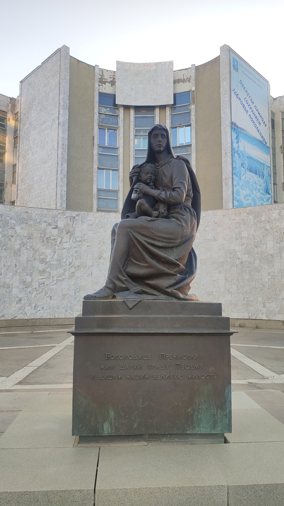 A bronze statue of the Virgin Mary holding the Christ child is situated in front of a building. The statue is on a dark bronze pedestal with an inscription in Cyrillic text. The building behind the statue is clad in white marble tiles and has several rows of windows. A large banner with a blue and white landscape image and text in Cyrillic is attached to the right side of the building. The sky is clear and pale. The statue and pedestal are centrally located on a paved area with geometric patterns. This is the monument to the Mother of God in Mirny, Russia.