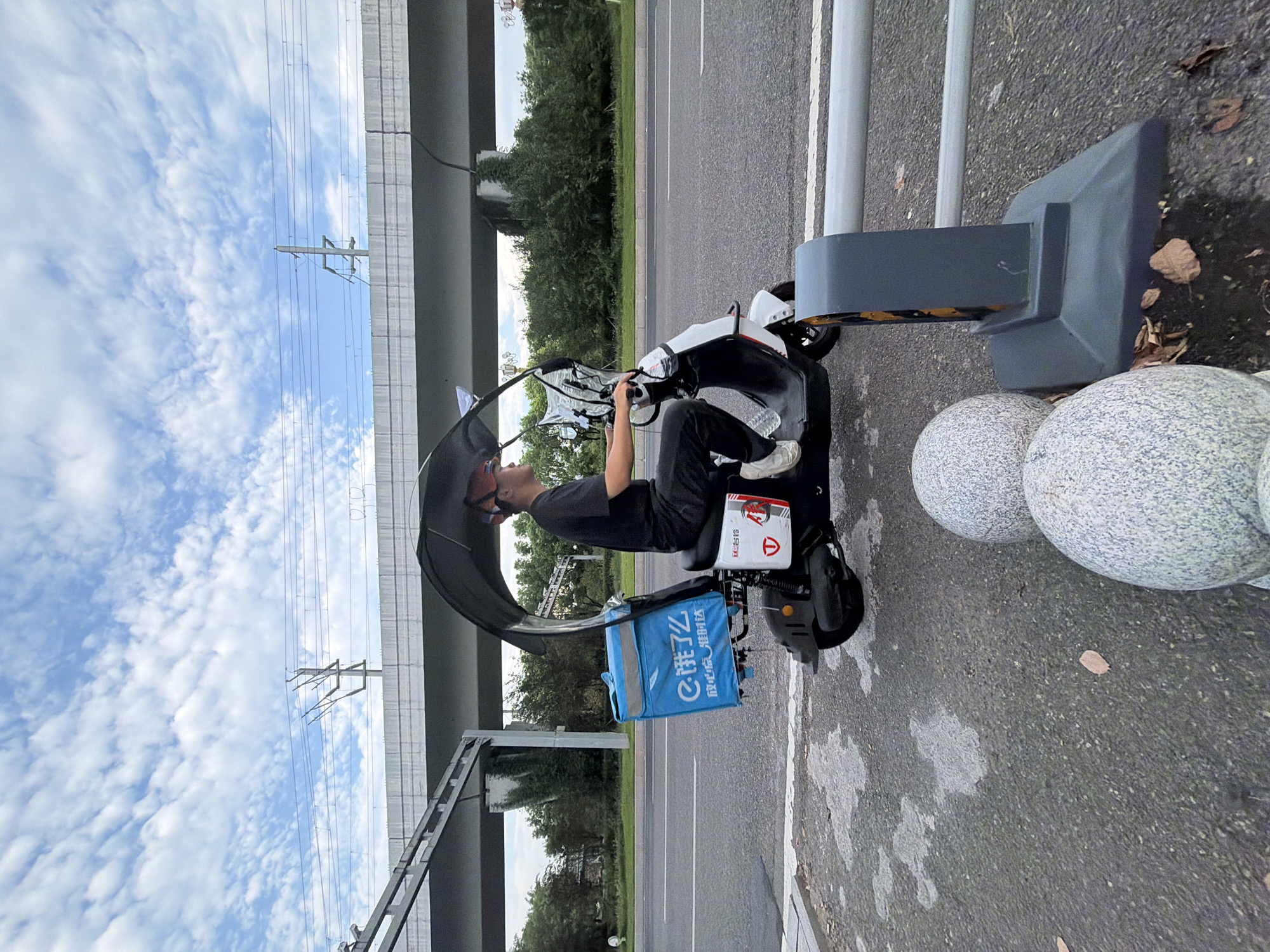 In Jiangjin, China, a delivery driver is captured riding a white electric scooter equipped with a blue insulated delivery bag. The driver, wearing a dark t-shirt and black pants, is seen from the side, looking forward. The scooter features a black canopy, providing shade. The background consists of a road, a pedestrian walkway, greenery, and an elevated highway structure with overhead power lines. The sky is blue with scattered white clouds. The scene is bordered by a grey guardrail and two spherical stone bollards.