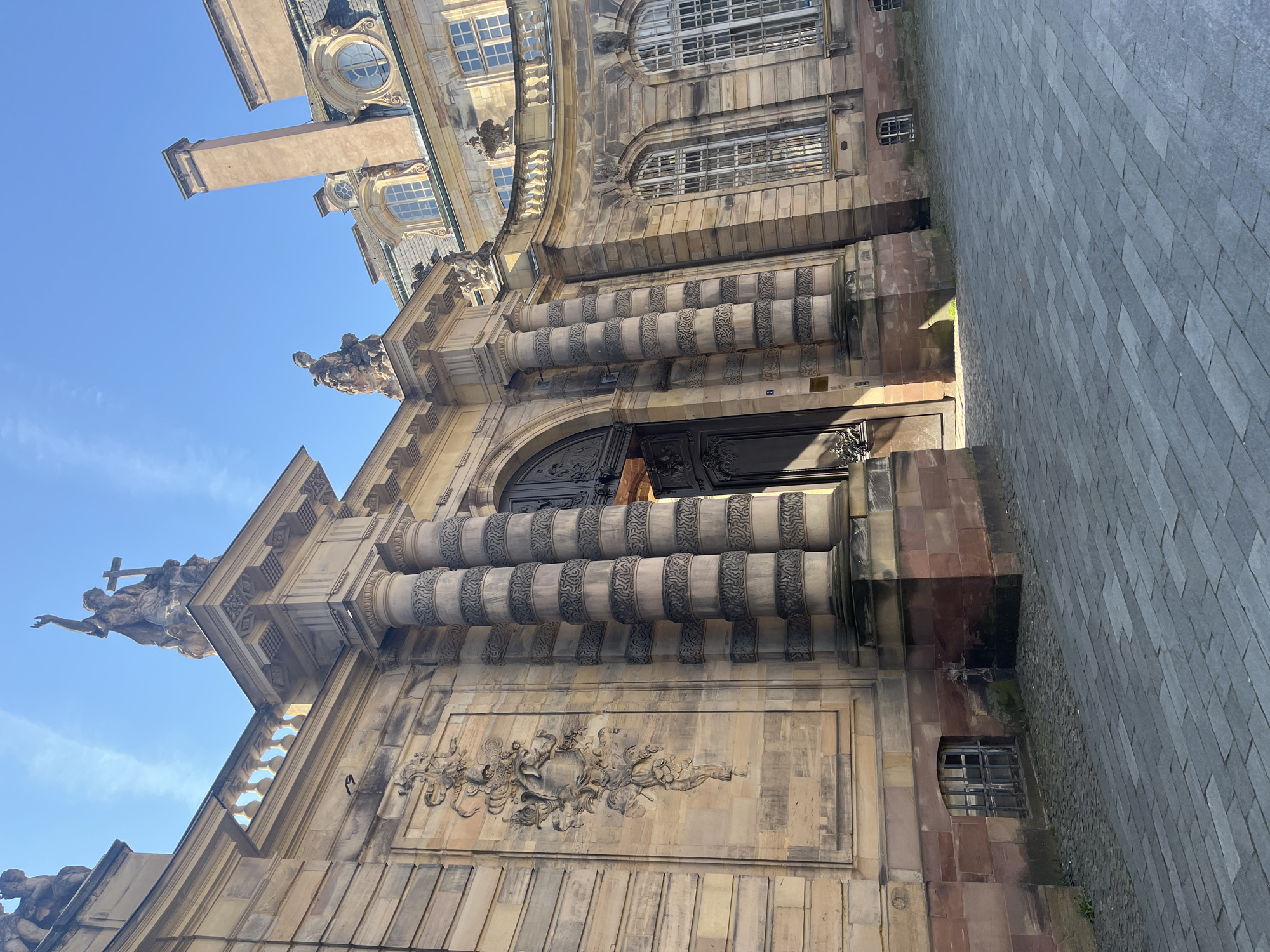 A low-angle shot captures the ornate facade of a grand historical building in Strasbourg, France, under a clear blue sky. The edifice is constructed primarily from light-colored stone, with sections of darker, reddish-brown stone adding textural contrast.
Dominating the lower-middle section is a large, dark, arched main entrance featuring intricately detailed doors, possibly made of wood or metal. Above this entrance, two prominent rows of decorative, banded columns or pilasters, adorned with swirling dark patterns, rise majestically, contributing to the building's elaborate grandeur. Below these columns, a finely carved rectangular relief sculpture stretches across the facade, depicting dynamic figures and organic motifs characteristic of Baroque or Rococo artistry.
Higher on the facade, various architectural elements include round oculus windows and rectangular windows fitted with metal grilles. The roofline is embellished with decorative cornices and balustrades, topped by several sculpted figures. On the upper-left side, a prominent statue of a robed figure holding a cross (possibly Christ or a saint) is silhouetted against the sky. A gargoyle-like sculpture also projects from a lower cornice, adding to the building's intricate details.
To the right of the building, a wide paved area, composed of light gray rectangular stones, suggests a public square or street. The clear, bright blue sky with no clouds indicates a sunny day, likely morning or early afternoon, as suggested by the shadows cast on the building and ground. A small white plaque bearing the number "24" is visible on the stone facade near the entrance. Another small, dark rectangular plaque is faintly visible on the lower reddish stone section, though its text is illegible. The overall impression is one of historical significance and meticulous architectural craftsmanship.