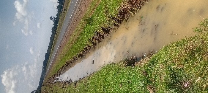 This image depicts a roadside environment located in the city of Heni Village, Kenya, under a sky with scattered white clouds and clear blue patches. A prominent, muddy water-filled drainage ditch occupies the left foreground, running diagonally towards the mid-right. The turbid, brown water within the ditch contains sparse vegetative debris. Its banks are covered with green grass. Immediately adjacent to the right of the ditch is a paved, two-lane asphalt road featuring a broken white center line, extending into the background. A light-colored vehicle is discernible in the distant portion of the road. Beyond the road, a dense line of green trees and foliage establishes the horizon.