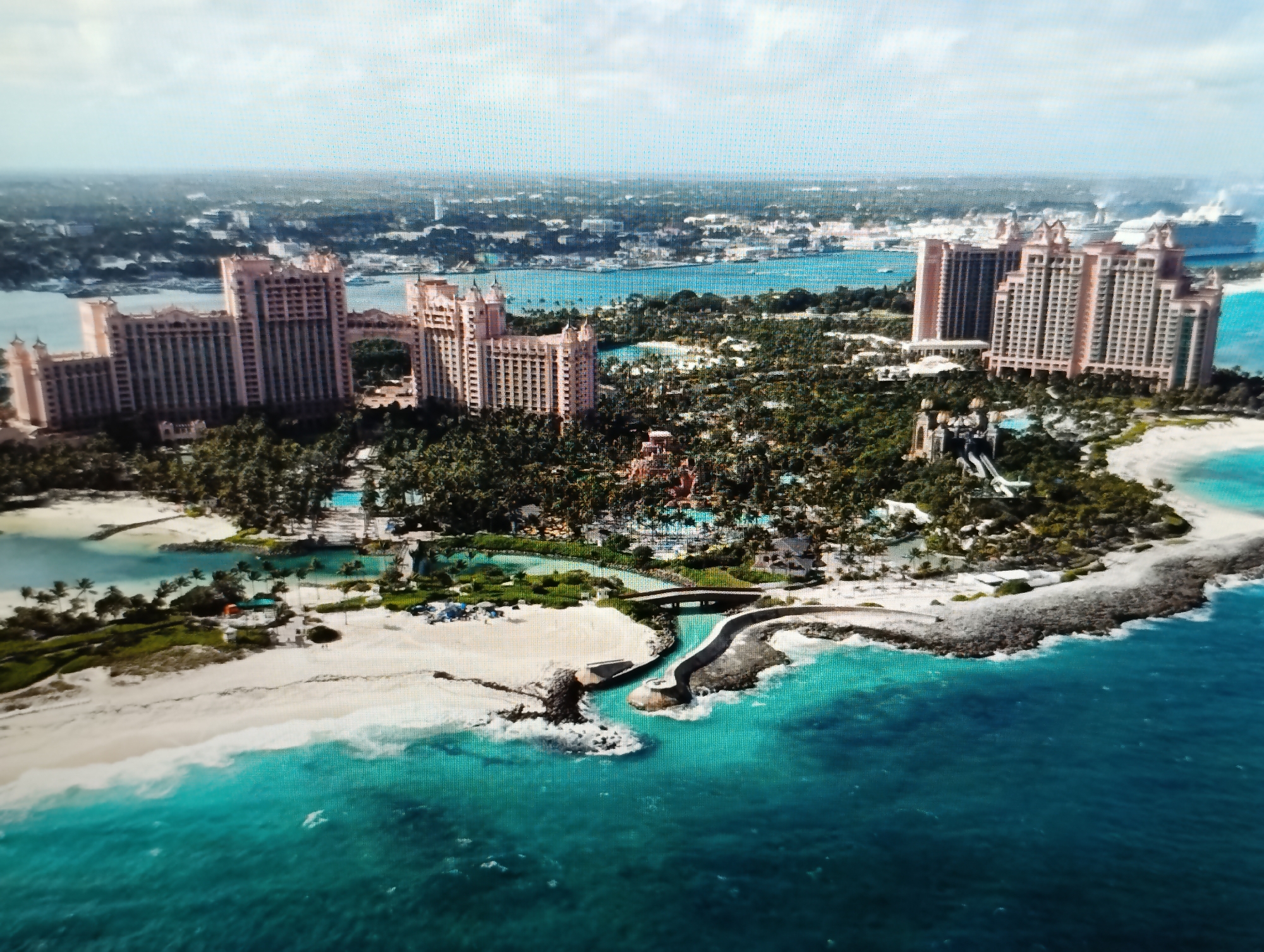 This is an aerial photograph of a resort complex on a tropical island. The resort features several large, pink-colored hotel buildings with distinctive architectural designs, including towers and ornate facades. Surrounding the hotels is a lush green landscape with numerous palm trees, indicating a warm climate. The resort also includes water features such as swimming pools and lagoons, some of which are visible with turquoise water. A white sand beach lines the edge of the island, meeting the clear, blue-green ocean. In the distance, a body of water connects to a larger expanse of ocean, and several cruise ships are visible near the coastline. The sky is partly cloudy, with a bright, hazy atmosphere. There is no visible indication of the city of Dubăsari town council, Moldova, in this image.