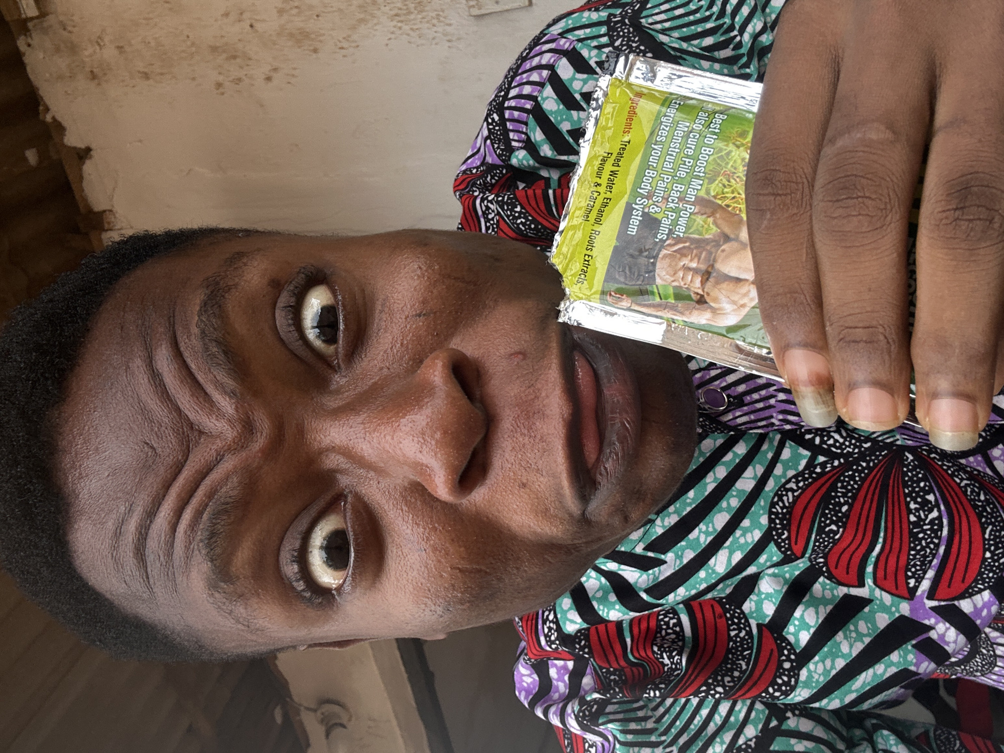 A person with dark skin, wearing a colorful shirt with red, green, black, and purple patterns, is holding a sachet of a product. The text on the sachet reads "Best to Boost Man Power, also cure Pile, Back Pains, Menstrual Pains & Energizes your Body System". The ingredients listed on the sachet are "Treated Water, Ethanol, Roots Extracts, Flavour & Caramel". The background seems to indicate an indoor location with a partly visible wall. The location is Unknown, Unknown.