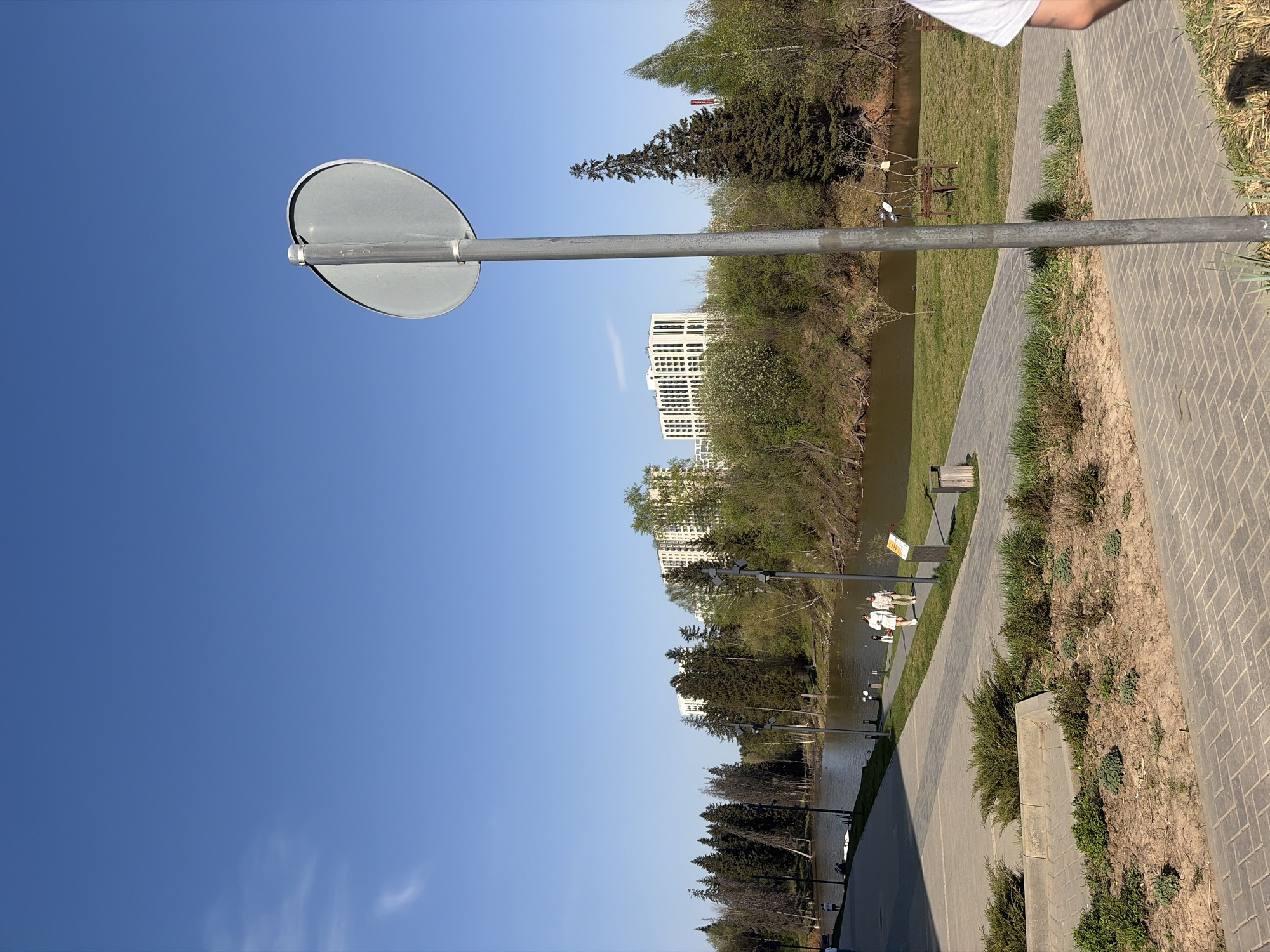 The image captures a view of Izhevsk, Russia. A grey road sign pole with a round, blank sign faces towards a clear blue sky, with a hint of cirrus clouds. To the right, a brick-paved walkway runs parallel to a river or body of water, bordered by grassy areas with shrubs and trees. The riverbank is lined with trees, and modern white buildings are visible in the background. People are present in the scene, walking along the walkway. The overall impression is a bright, sunny day in a park setting within the city.
