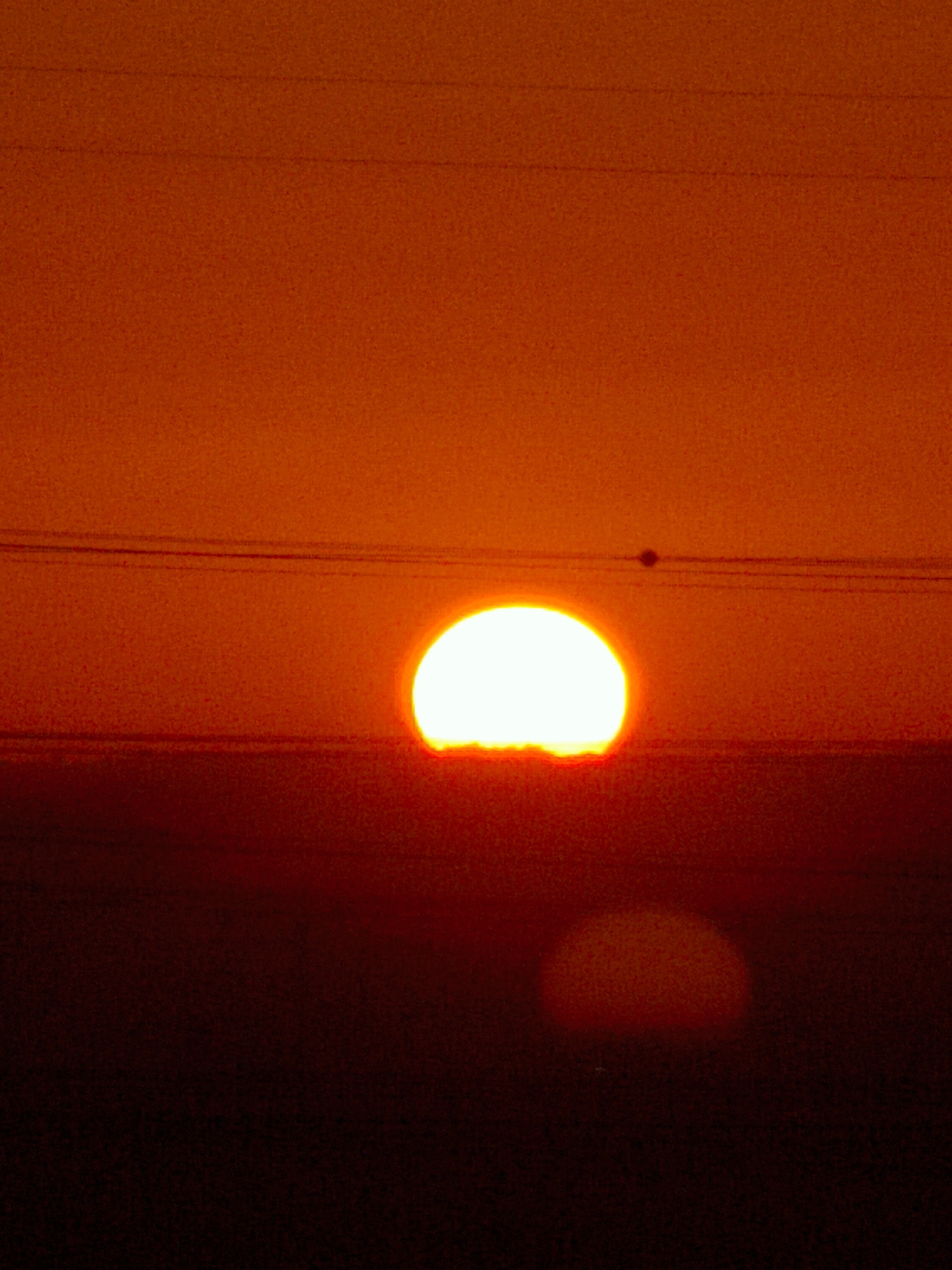 The image captures a sunset over Be'er-Sheva, Israel. A bright, white sun, appearing as a semicircle, is positioned low on the horizon, partially obscured by a dark silhouette that suggests the ground or a distant landscape. Two thin, dark horizontal lines, likely power lines, stretch across the upper portion of the sky, with one line passing just above the sun. A small, dark circular object, possibly a bird or an insect, is visible in the sky between the power lines and the sun. The sky is a gradient of deep orange and red, transitioning to a darker red and black towards the bottom of the frame, indicative of the fading daylight. The overall scene is dominated by warm, intense colors associated with a sunset.