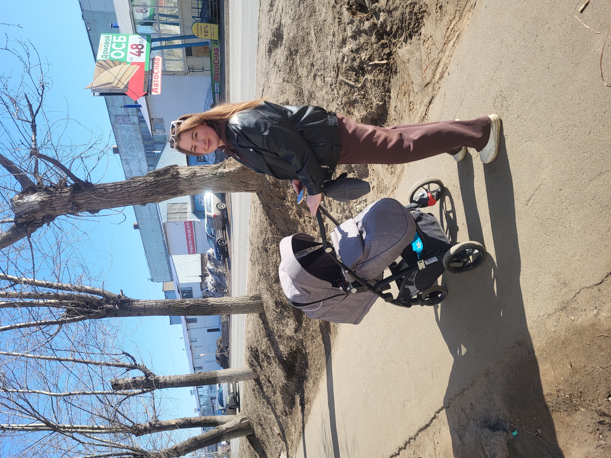 A woman with long, light-brown hair is standing on a sidewalk next to a tree with bare branches, pushing a baby stroller. The woman is wearing sunglasses on her head, a black leather jacket, and brown pants, and she is looking at the camera. The stroller is gray and black.
In the background, there is a building with signs written in Russian, including a sign that says "Домовой ОСБ" and "АВТОСНАБ," along with an indication of the price of 48 rubles. There are also cars parked along the street.
The scene appears to be a sunny day in the city of Chaikovsky, Russia, based on the Russian text in the signs.
