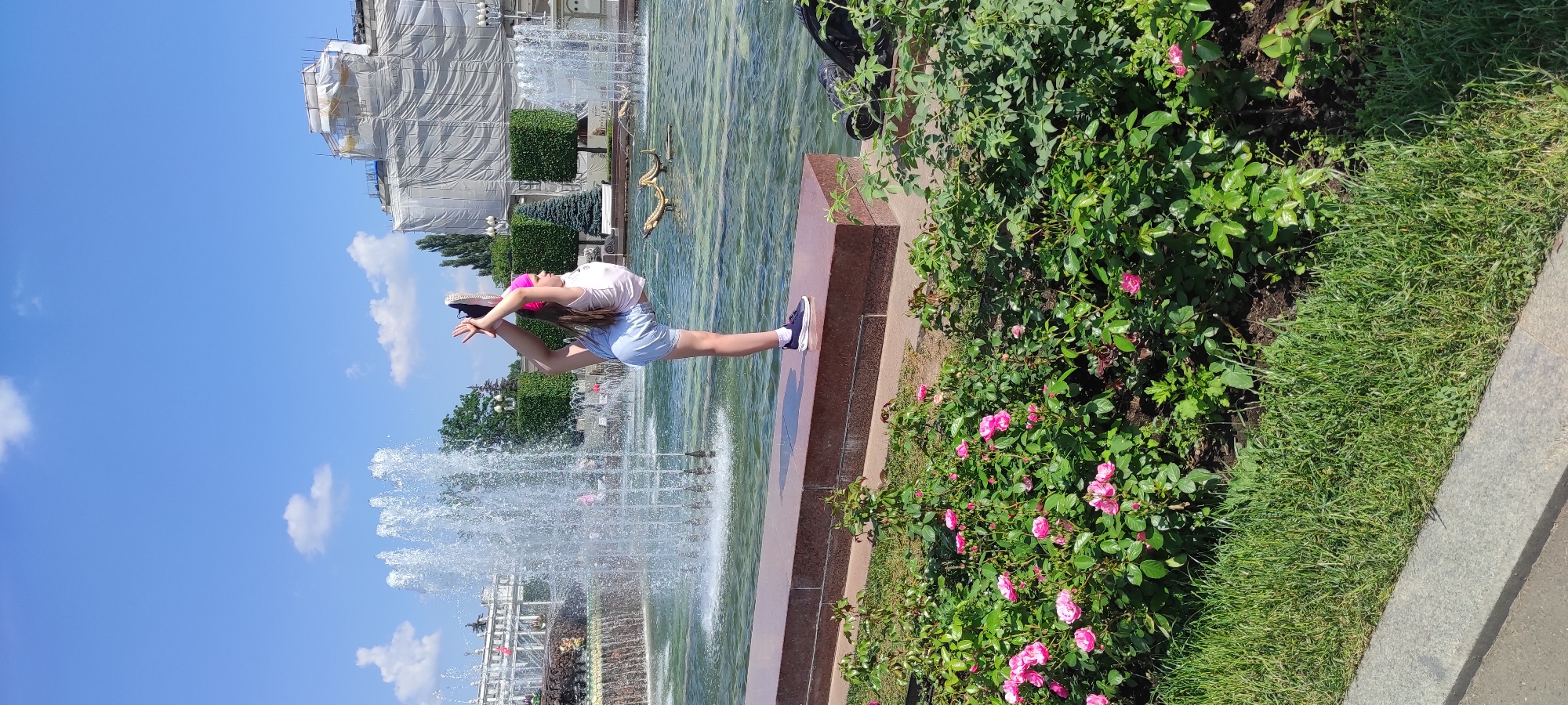 In Moscow, Russia, a young girl in a white t-shirt, blue shorts, and a pink headband performs an extreme backbend pose with her right foot held high behind her head. She is standing on a raised, red-brown stone ledge in front of a large, multi-tiered fountain. The fountain's water sprays upwards and outwards, creating a shimmering effect. Behind the fountain, a large building is partially covered in white scaffolding and tarps, suggesting ongoing construction or renovation. The surrounding area features manicured green bushes and trees, with a bed of pink roses in the foreground. The sky is bright blue with a few scattered white clouds. The overall scene suggests a sunny day in a public park or plaza.