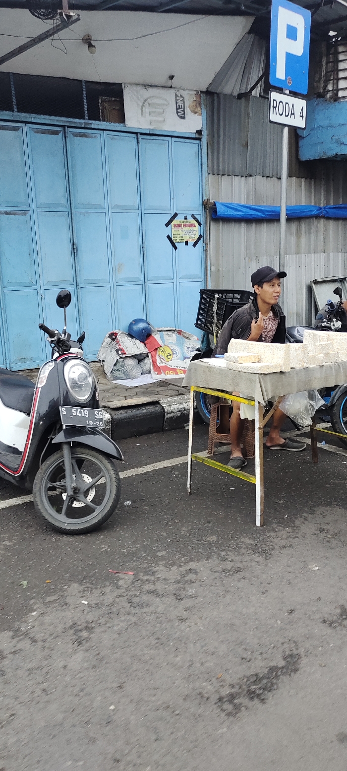 The image shows a street scene in Mojokerto, Indonesia, located at coordinates -7.467 latitude and 112.438 longitude. A man in a black baseball cap and a pink and black patterned shirt sits at a table, his legs crossed, holding a white object in his left hand. The table is covered in a grey cloth and has a stack of white objects on it. There is a black and white scooter parked on the left side of the image. The scooter has a license plate that reads "S 5419 SO 10-26". There is a blue sign with a white letter "P" and the words "RODA 4" in white on it. There is a blue tarp on the building in the background and a blue corrugated metal wall behind it. The man is sitting on a yellow stool, and there is a yellow-painted curb separating the street from the sidewalk. The ground in the image is a grey, paved road, and there is a black curb in the foreground. The building in the background is made of concrete and has a blue paint job. There are several wires hanging from the building's ceiling, and there is a sign that reads "NEW" in the background. The image is taken during the daytime, and there is no sunlight shining directly on the scene.