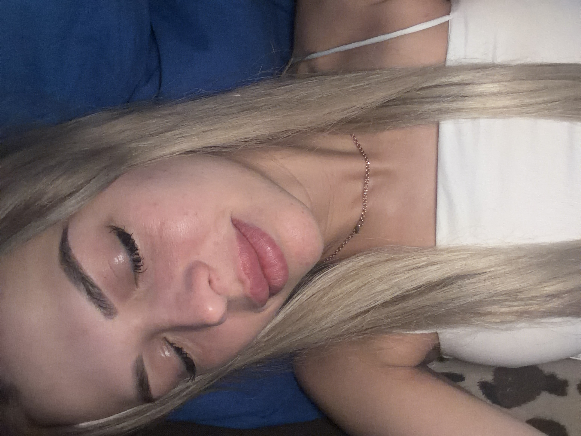 A close-up, head-and-shoulders portrait of a young woman with blonde hair is shown from the chest up. She is lying down with her eyes closed and appears to be wearing a white strapless top and a thin, sparkly necklace. The background is a dark blue fabric, possibly a pillow or blanket. No specific location in Kyiv, Ukraine, can be identified from the image.