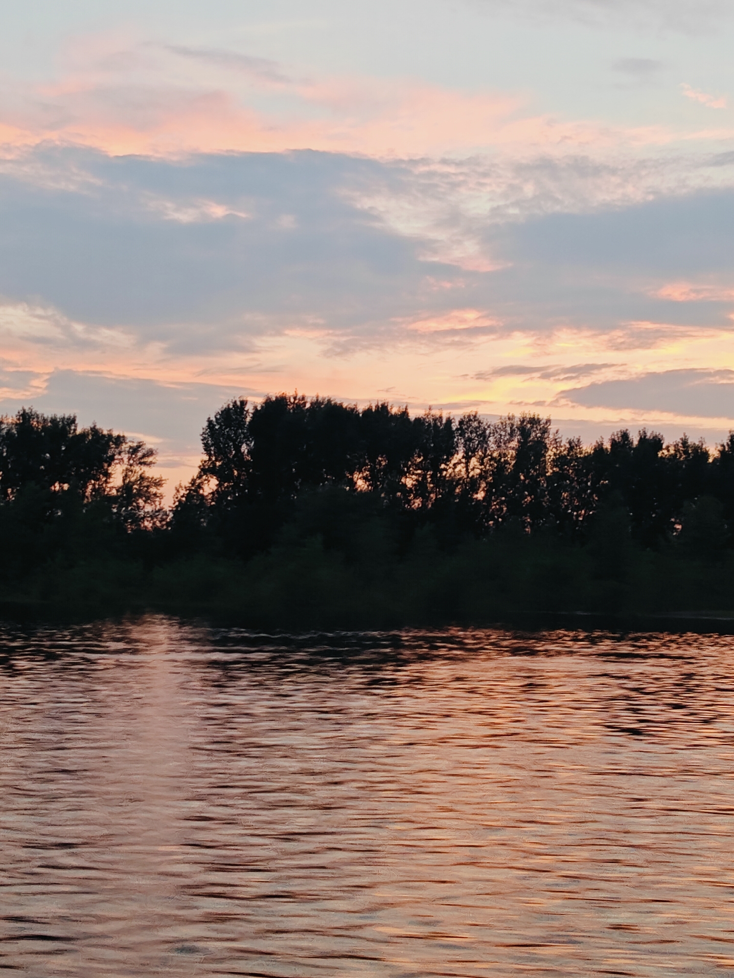 The image shows a serene natural scene at dusk, featuring a wide body of water in the foreground and a treeline against a soft, pastel-colored sky. The water is calm, with gentle ripples reflecting the warm hues of the sunset. The sky is a blend of pale pinks, oranges, and blues, with scattered, wispy clouds. A dense line of dark green trees forms a silhouette along the horizon, partially obscuring the setting sun. The overall impression is one of tranquility and natural beauty. The location is identified as Noviy, Russia, suggesting a rural or riverside setting.