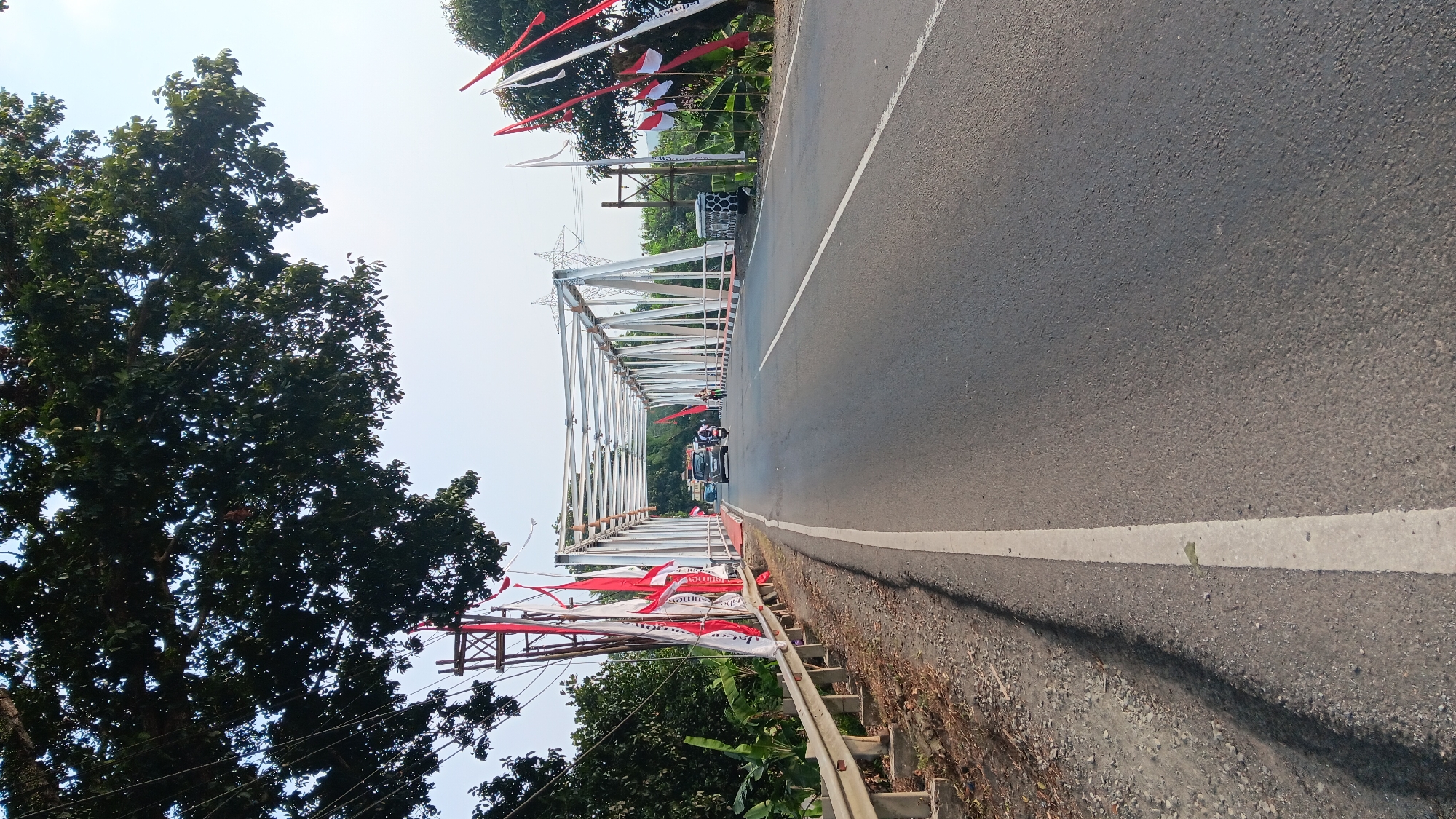 The image captures a view of a bridge crossing a road in Tanjungsari, Indonesia, under a bright, hazy sky. The bridge is constructed with a white metal truss structure and is decorated with numerous red and white flags, likely representing the Indonesian flag. A paved road runs across the bridge, with a white line marking the lane. Vehicles, including a dark-colored car and a motorbike, are visible on the road, suggesting active traffic. On the left side of the road leading up to the bridge, a metal guardrail runs along the edge, and tall trees with dense green foliage overhang the scene. The environment appears to be semi-rural, with vegetation visible on either side of the road and in the background. The lighting suggests it is daytime.