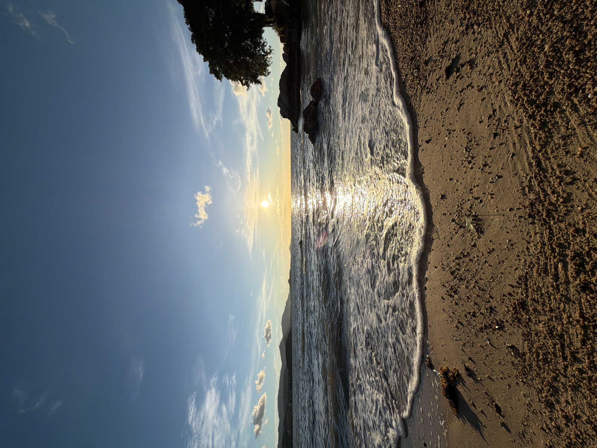 The image depicts a serene beach scene at sunset in Baan Bo Phut, Thailand. The sun is low on the horizon, casting a warm, golden light that reflects shimmering on the gentle waves. The sky is a clear, vibrant blue, transitioning to hues of orange and yellow near the sun. Wispy white clouds are scattered across the sky, adding a touch of softness to the scene.
The foreground is dominated by a sandy beach, with the wet sand at the water's edge appearing darker and more compact. Small pebbles and bits of seaweed are visible on the sand. The ocean is calm, with small waves rolling in and leaving a frothy white line of foam along the shore. In the background, distant hills or mountains form a silhouetted outline against the bright sky. To the left, the dark silhouette of a leafy tree and some rocks are visible near the water's edge.
The overall mood of the image is peaceful and tranquil, evoking a sense of calm and relaxation associated with a beach at the end of the day. The natural beauty of the sunset over the ocean creates a visually appealing and inviting atmosphere. No text is visible in the image.