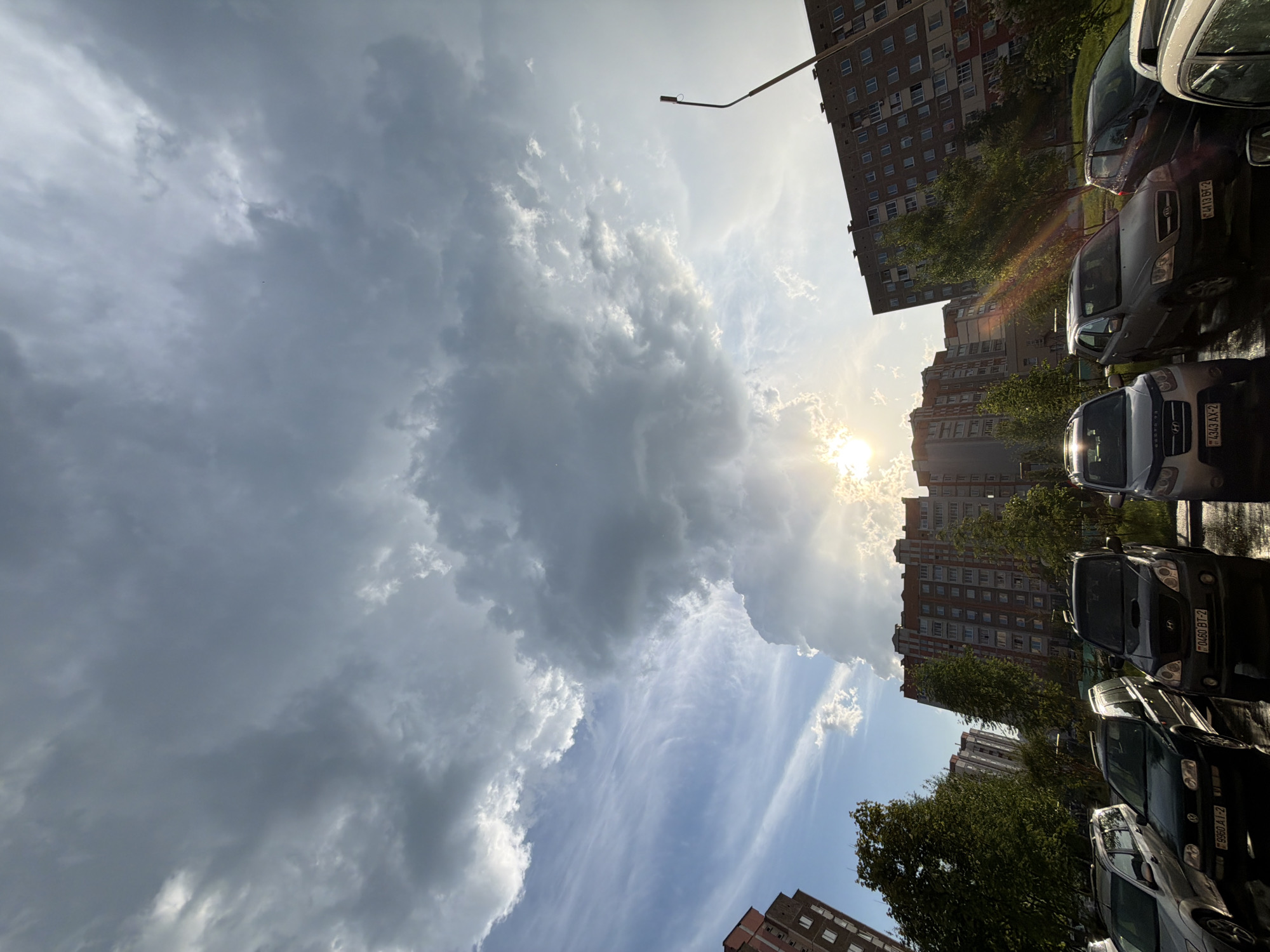 The image shows a row of parked cars in front of tall apartment buildings under a cloudy sky with the sun shining through. The location is in an urban area, possibly a residential neighborhood, although the specific city is unknown. The cars are parked close together, suggesting a densely populated area. The buildings are multi-story and appear to be residential apartments. The sky is filled with clouds, with patches of blue visible between them. The sun is partially obscured by the clouds, creating a bright glare in the center of the image. A streetlight can be seen near the buildings. The image is taken from a low angle, looking up towards the sky.