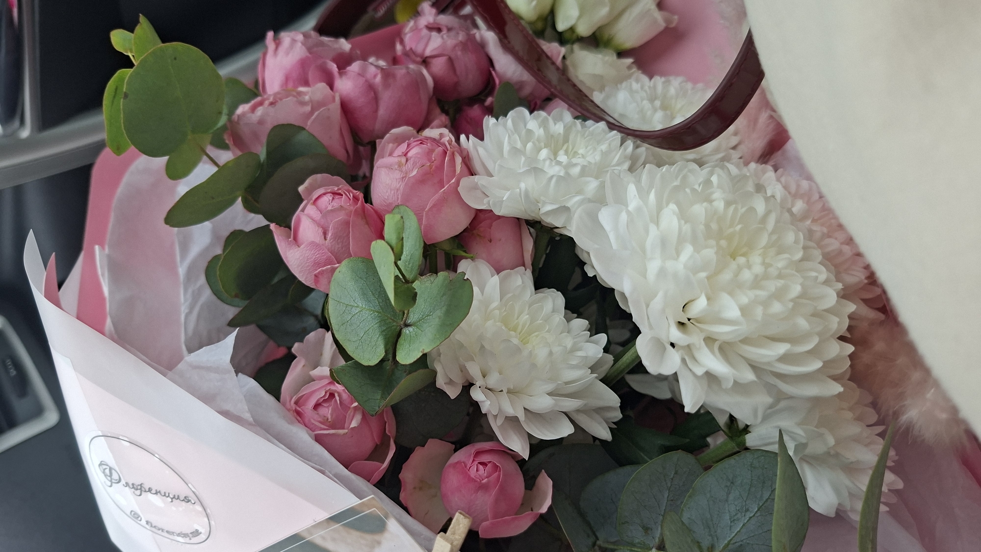 A bouquet of flowers is presented in a wrapping. The flowers include pink roses and white chrysanthemums. The bouquet also features sprigs of green eucalyptus leaves. The wrapping is predominantly white and pink, with text that appears to be in a Cyrillic script on the lower left. The setting is indoors, with a portion of a car interior visible. Location in the city of Дзяржынскі сельскі Савет, Belarus is not discernible from the image.