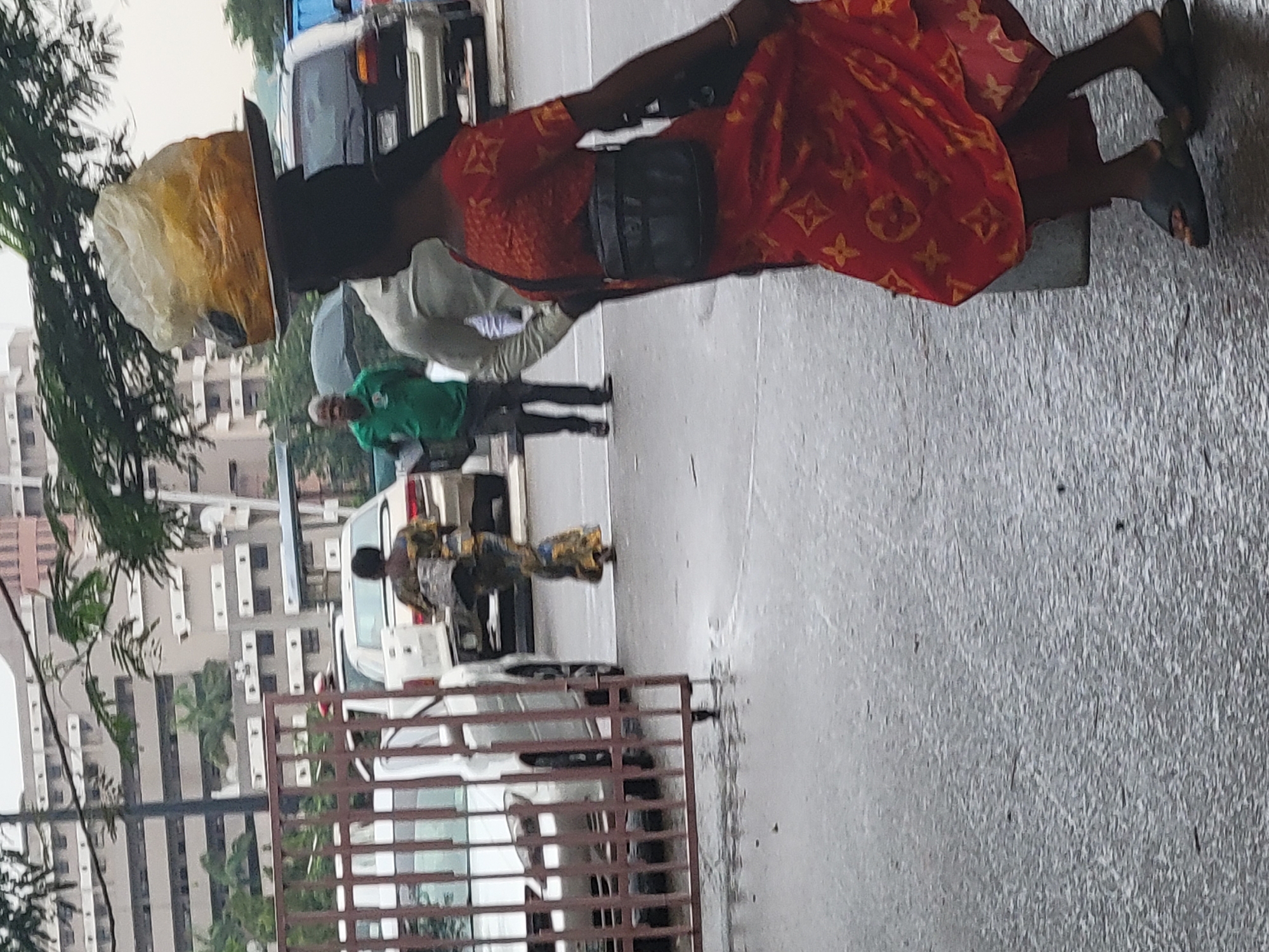 A street scene in Abuja, Nigeria. A woman in an orange dress with gold patterns walks toward the right side of the frame, carrying an item on her head. A black bag is slung over her shoulder. In the background, two people are walking, one wearing a green shirt. Cars are parked along the street. A metal gate is in the foreground. Buildings are visible in the distance. The street appears wet, suggesting recent rain.