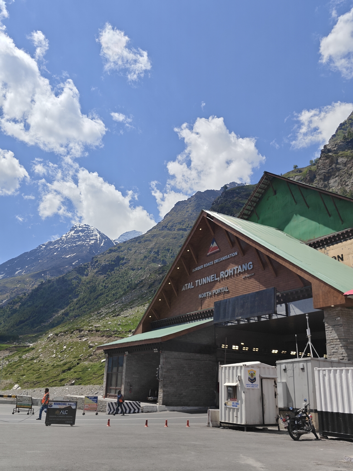 In an outdoor scene at the north portal of the Atal Tunnel, Rohtang, in the mountains of India, a structure with a wooden facade and a green roof stands prominently. The facade displays text that reads "BORDER ROADS ORGANISATION ATAL TUNNEL, ROHTANG NORTH PORTAL". Two men are present on the paved area in front of the structure. One man, wearing an orange vest, pushes a cart with a sign that says "THE BULEVAR" and "HIMACHAL POLYDE ALC". Another man, also in an orange vest, walks towards the structure. A motorcycle is parked to the right of a white portable building. The environment features a steep, green, and rocky mountainside in the background under a bright blue sky with scattered white clouds. Several orange traffic cones are placed on the pavement.