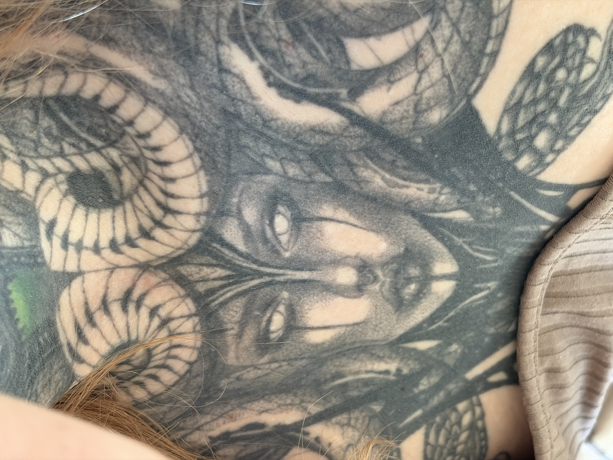 A close-up photograph shows a tattoo of a face with snakes for hair on a person's back or shoulder. The tattoo is done in black and gray ink. A portion of the person's hair is visible in the lower left corner. A beige striped garment is visible on the right edge of the image. There is no information about the location of the image within the city of Chaikovsky, Russia.