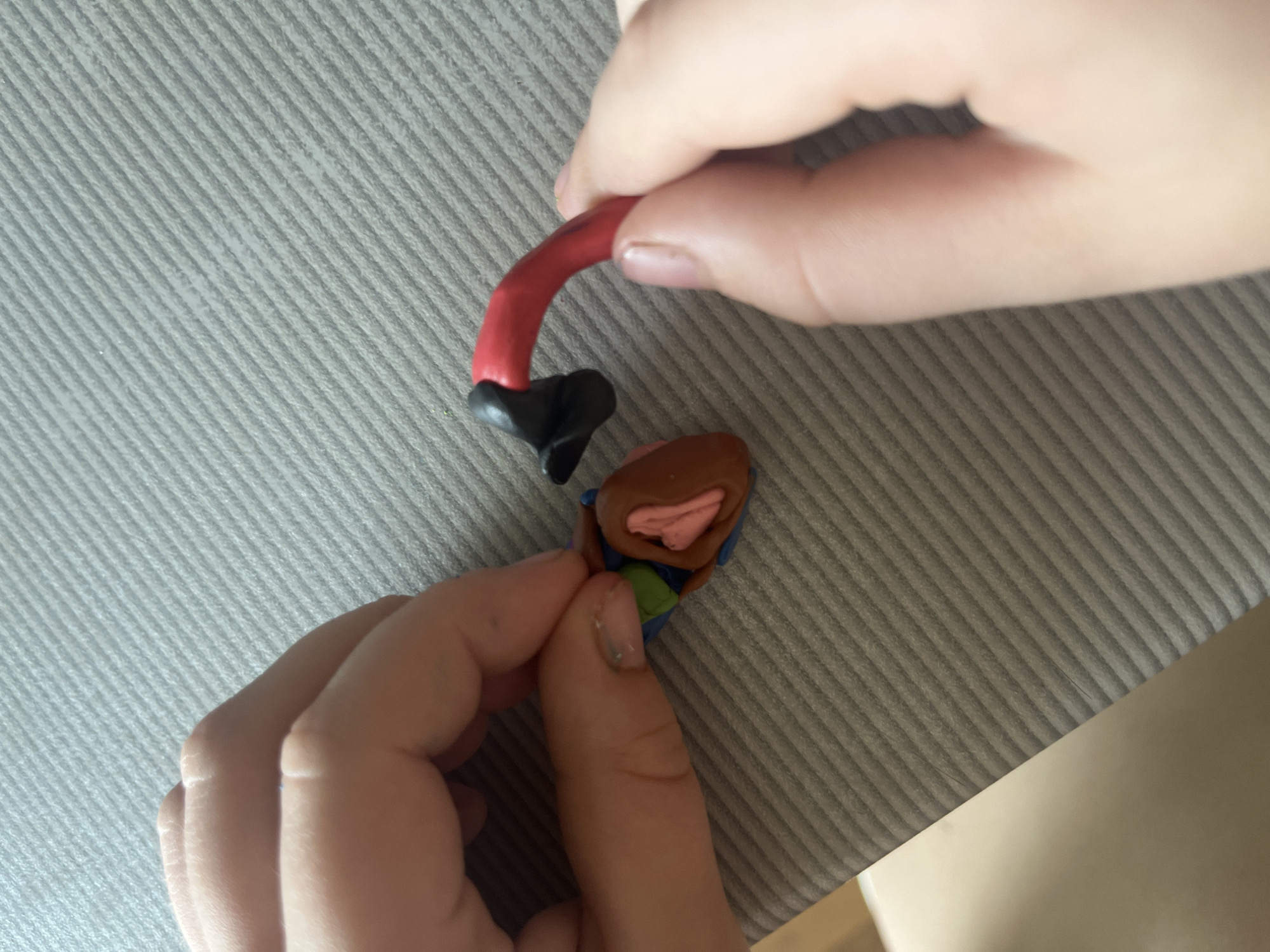 The image shows a close-up of a person's hands shaping a heart-like object made of clay. The clay heart is brown with pink and green accents. A red and black piece of clay, also shaped like a limb or appendage, is being attached to the heart. The background is a gray, textured surface, possibly a table or mat. The image does not contain any information that can be used to determine a location in Sochi, Russia.