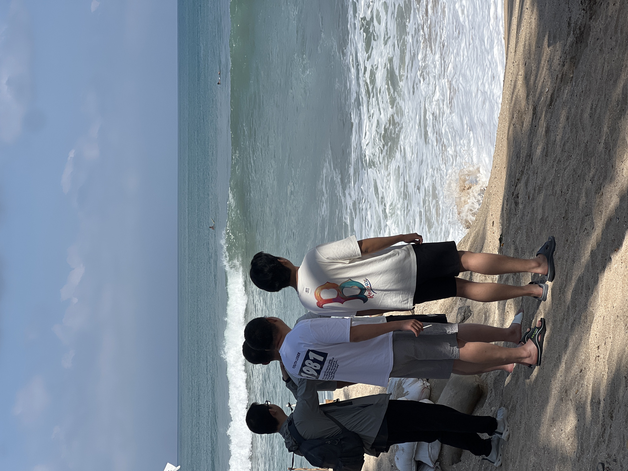 Four men stand on a sandy beach in Kuta, Indonesia, looking out at the ocean. The sea is a clear turquoise color, with gentle waves breaking on the shore. There are two surfers in the water, one further out than the other. The men are casually dressed, and the sky is a bright blue with a few scattered clouds. The foreground of the image shows sand and some sandbags, suggesting that this area may be prone to erosion or flooding. The men appear to be observing the surfers or the ocean, and their attention is focused outwards.