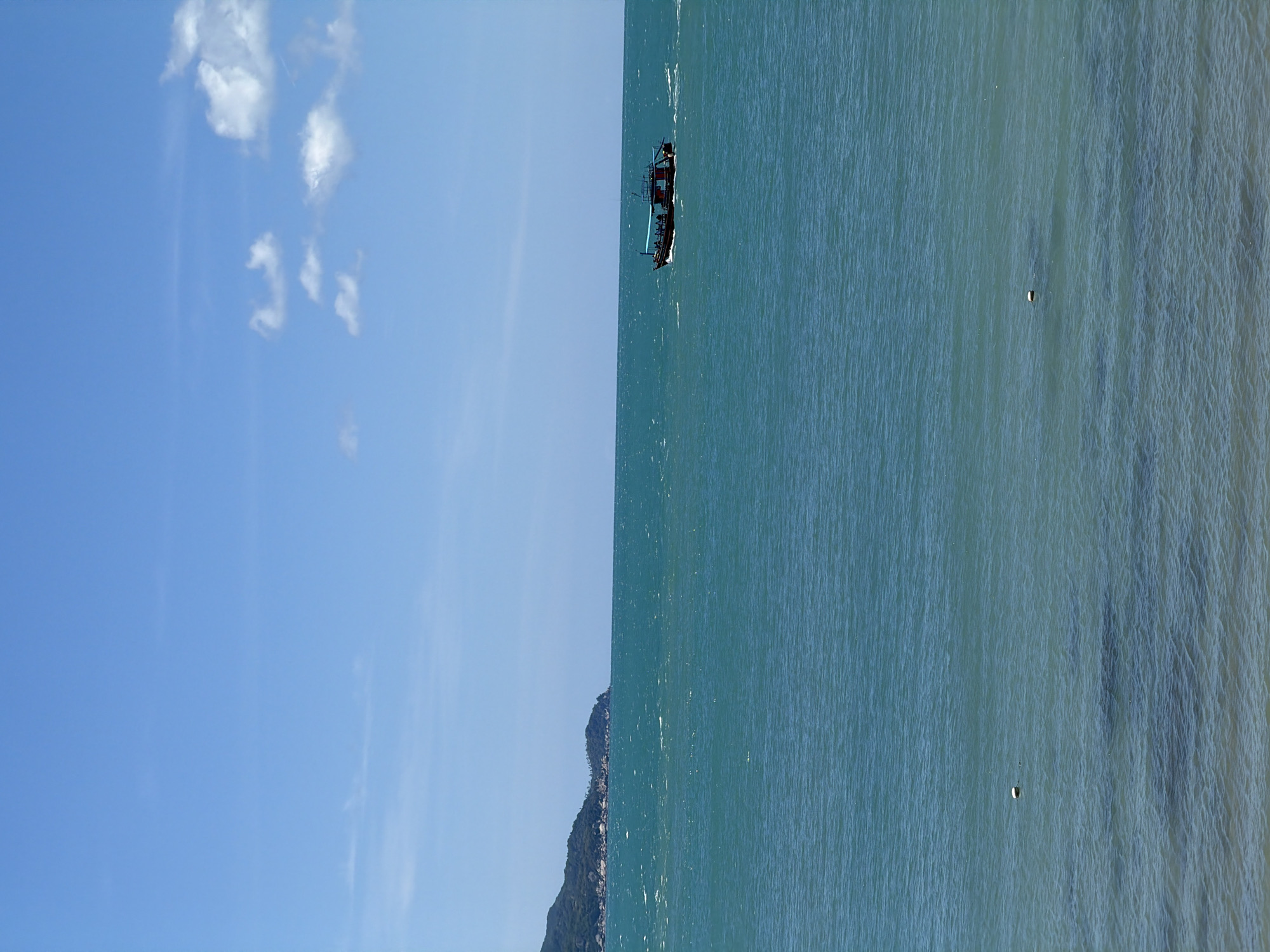 The image, taken at Baan Plai Laem, Thailand, captures a serene coastal scene. The composition is bisected by a clear horizon line, with the left side dominated by a vast, clear blue sky dotted with wispy white clouds, suggesting a bright, sunny day. On the right, a tranquil, turquoise sea stretches out, showing subtle ripples and a few distant buoys, indicating calm conditions.
A small, dark boat, likely a fishing vessel with a rudimentary canopy, is the central object of interest on the water. It appears to be moving, leaving a faint wake behind it. The boat is positioned mid-frame, drawing the viewer's eye towards its solitary presence on the expansive ocean. In the distance, a rocky, tree-covered island juts out from the sea on the left side of the image, adding a touch of dramatic landscape to the otherwise open view.
The overall mood is peaceful and contemplative, evoking the quiet beauty of a tropical coastal environment. The time of day is likely midday or early afternoon, given the bright sunlight and clear skies. There is no visible text in the image.