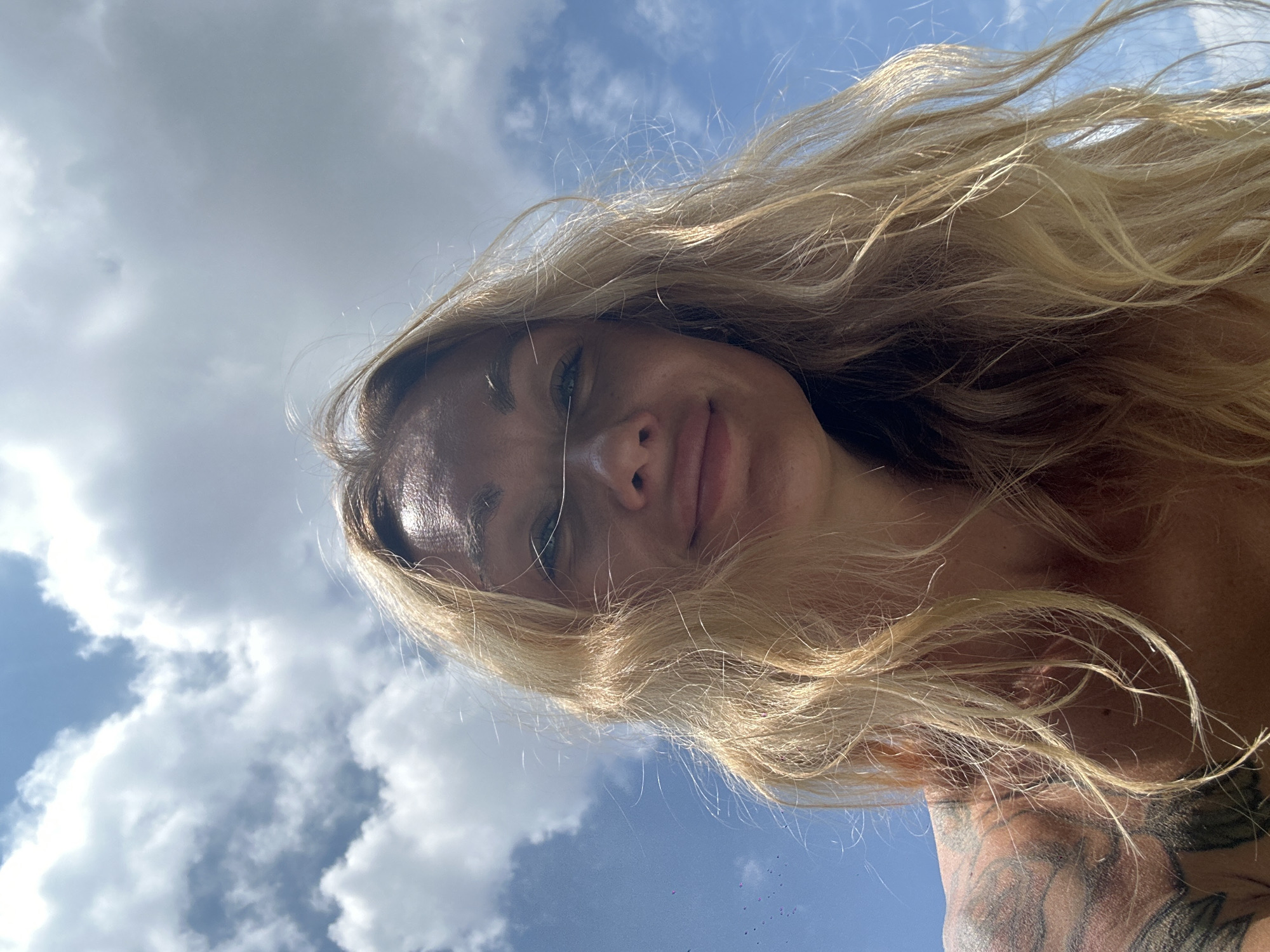 An extreme low-angle, outdoor, waist-up selfie of a fair-skinned, blonde woman is captured against a backdrop of a bright blue sky filled with fluffy white clouds. The woman's hair is long, wavy, and blonde, with strands blowing in the wind. Her face is partially obscured by her hair, but her eyes are visible, looking towards the camera. She has a slight smile on her lips. Her shoulder and upper chest are visible, revealing a tattoo on her left shoulder. The scene appears to be taken on a sunny day, possibly in a coastal area due to the clear sky and open space. No specific landmarks or details identifiable to Mũi Né, Vietnam, are present in the image.